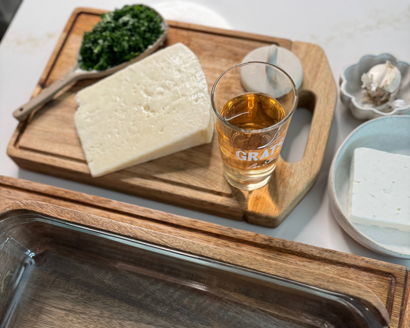 A wooden cutting board with a block of white cheese, a glass of amber liquid, and a spoonful of chopped greens. Nearby are a dish of feta cheese and a small bowl with garlic. A glass baking dish is in the foreground.