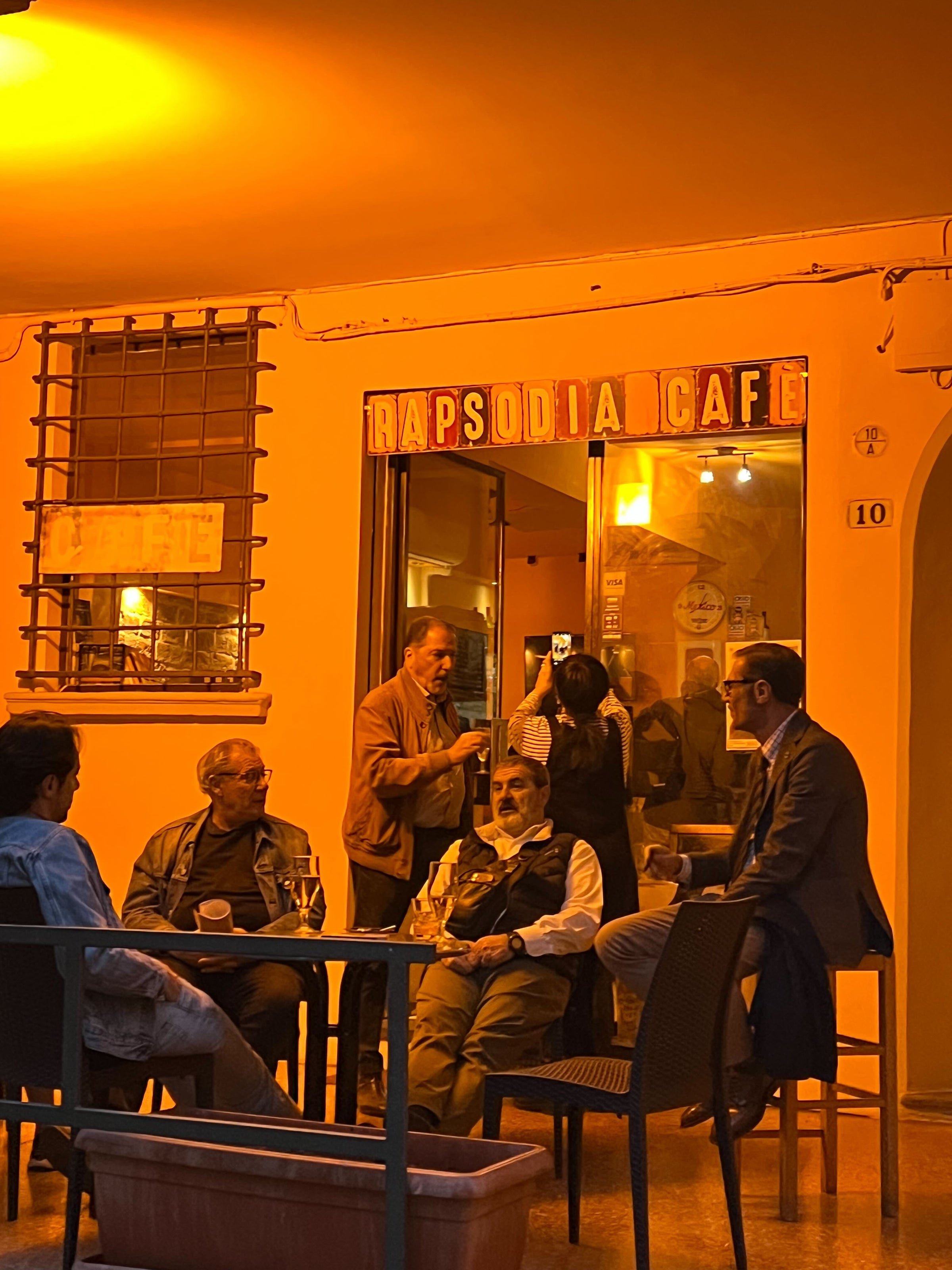 A group of people sit and stand outside Rapsodia Café at night, illuminated by warm yellow-orange light. Some are talking while others are taking photos near the entrance under a sign reading RAPSODIA CAFE.