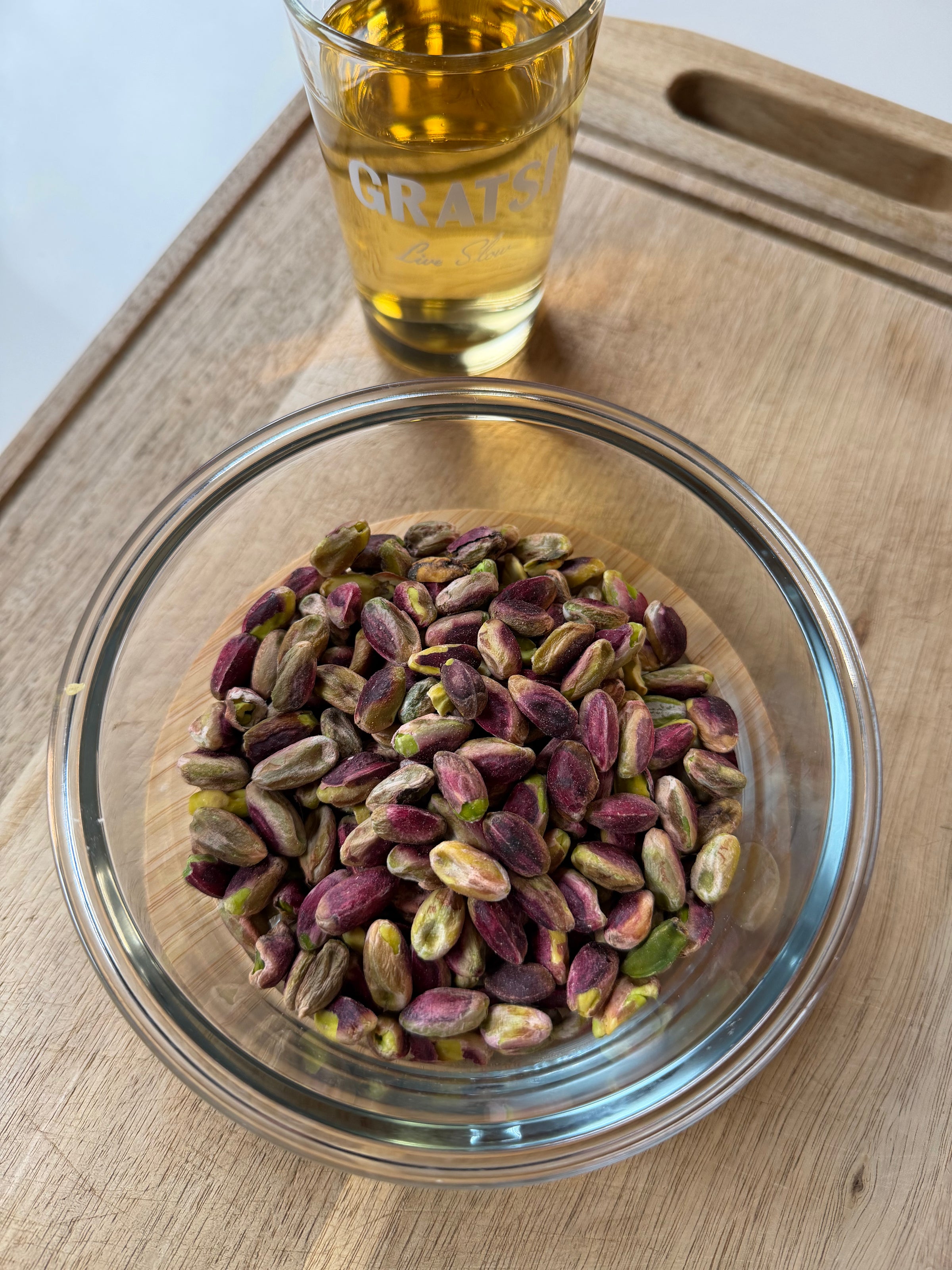 A glass bowl filled with shelled pistachios sits on a wooden cutting board, next to a glass of golden-colored liquid.