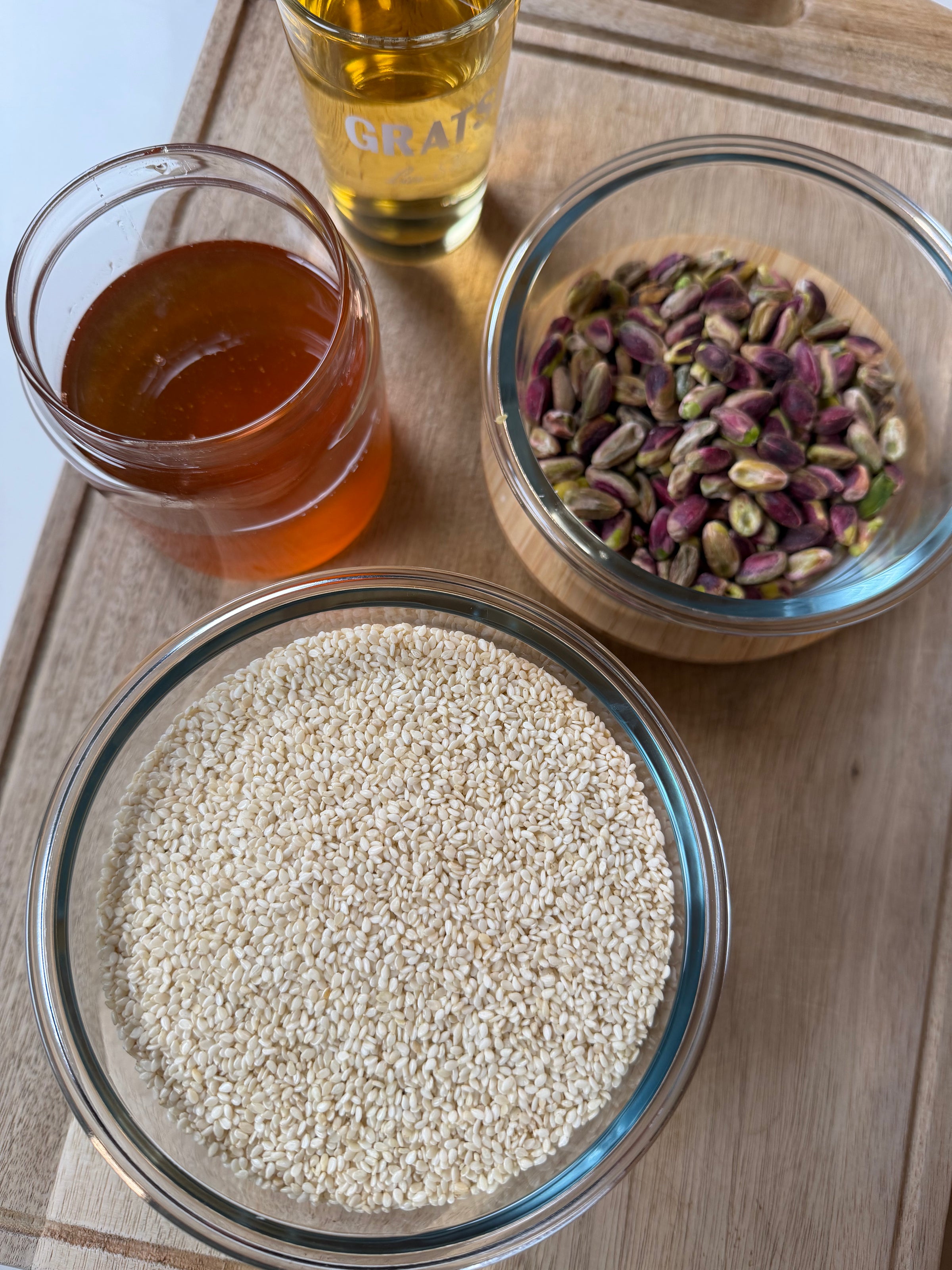 Four glass bowls on a wooden surface containing honey, pistachios, sesame seeds, and a small glass of clear golden liquid, possibly oil or syrup, arranged neatly.