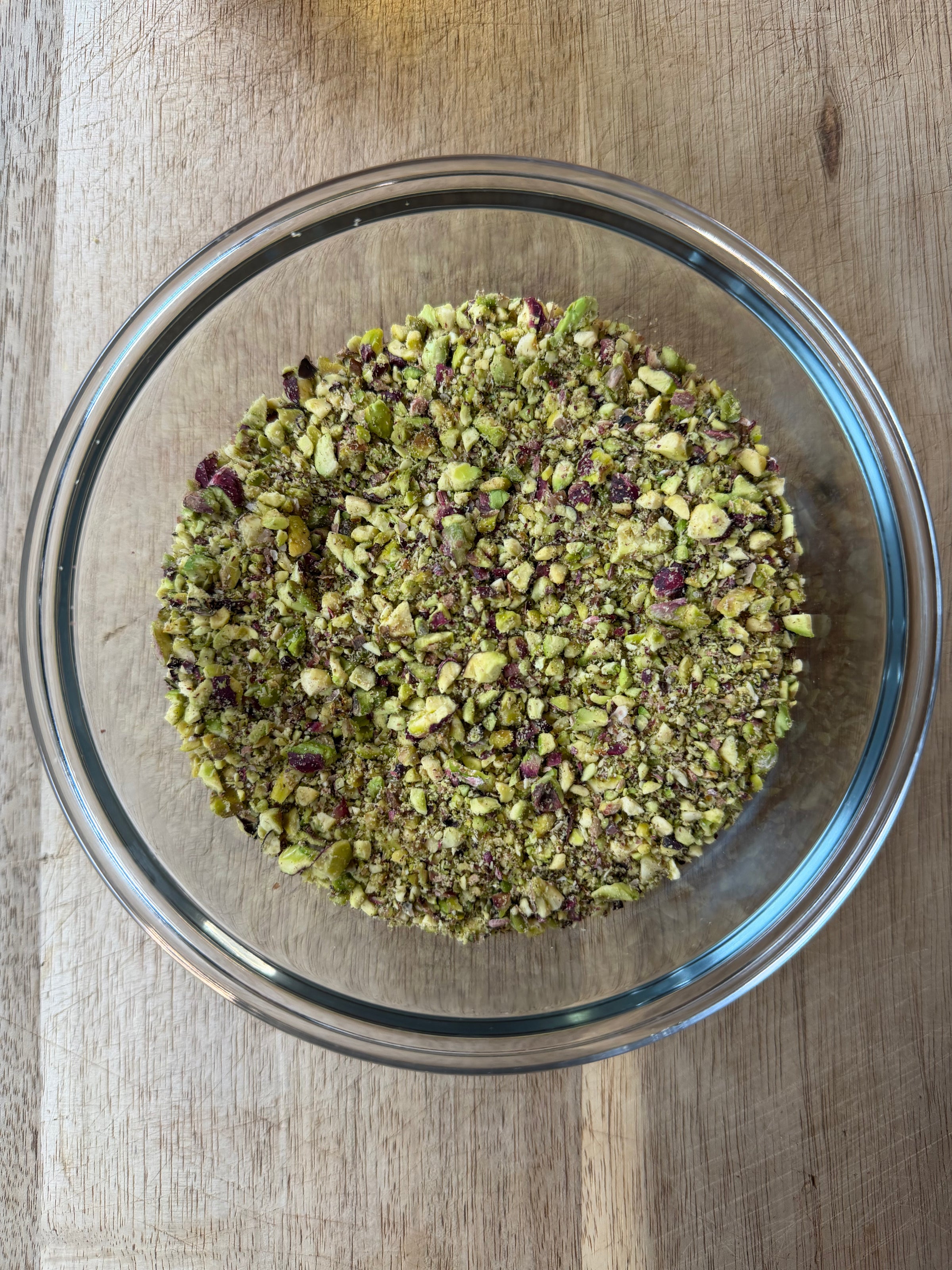 A glass bowl filled with finely chopped pistachios sits on a wooden surface. The nuts appear green with hints of purple and tan.