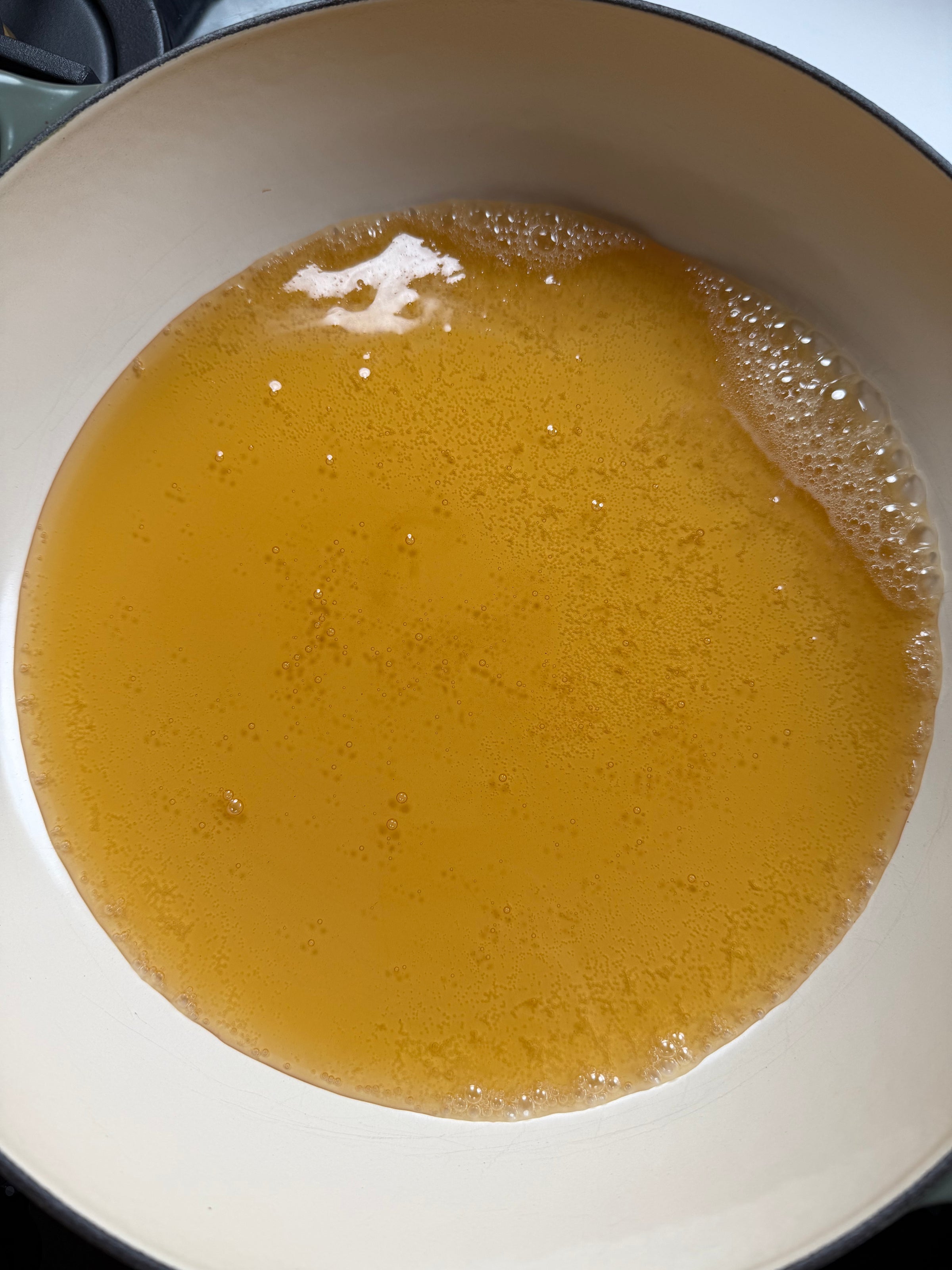 A close-up of a white pot filled with a warm, golden-brown liquid, likely broth or stock, with small bubbles forming on the surface.