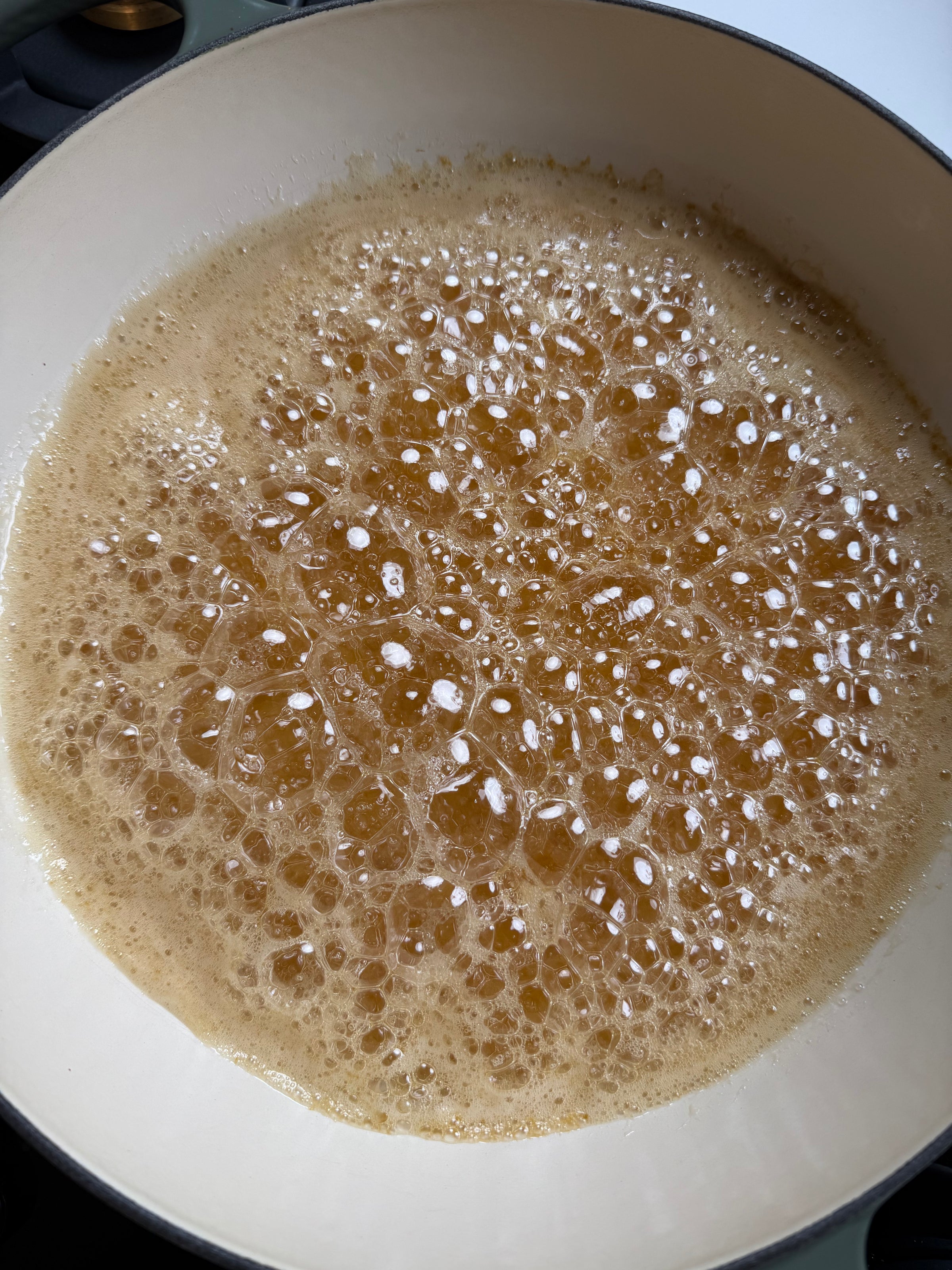 A close-up of golden-brown bubbling caramel cooking in a white enamel pan on a stovetop.