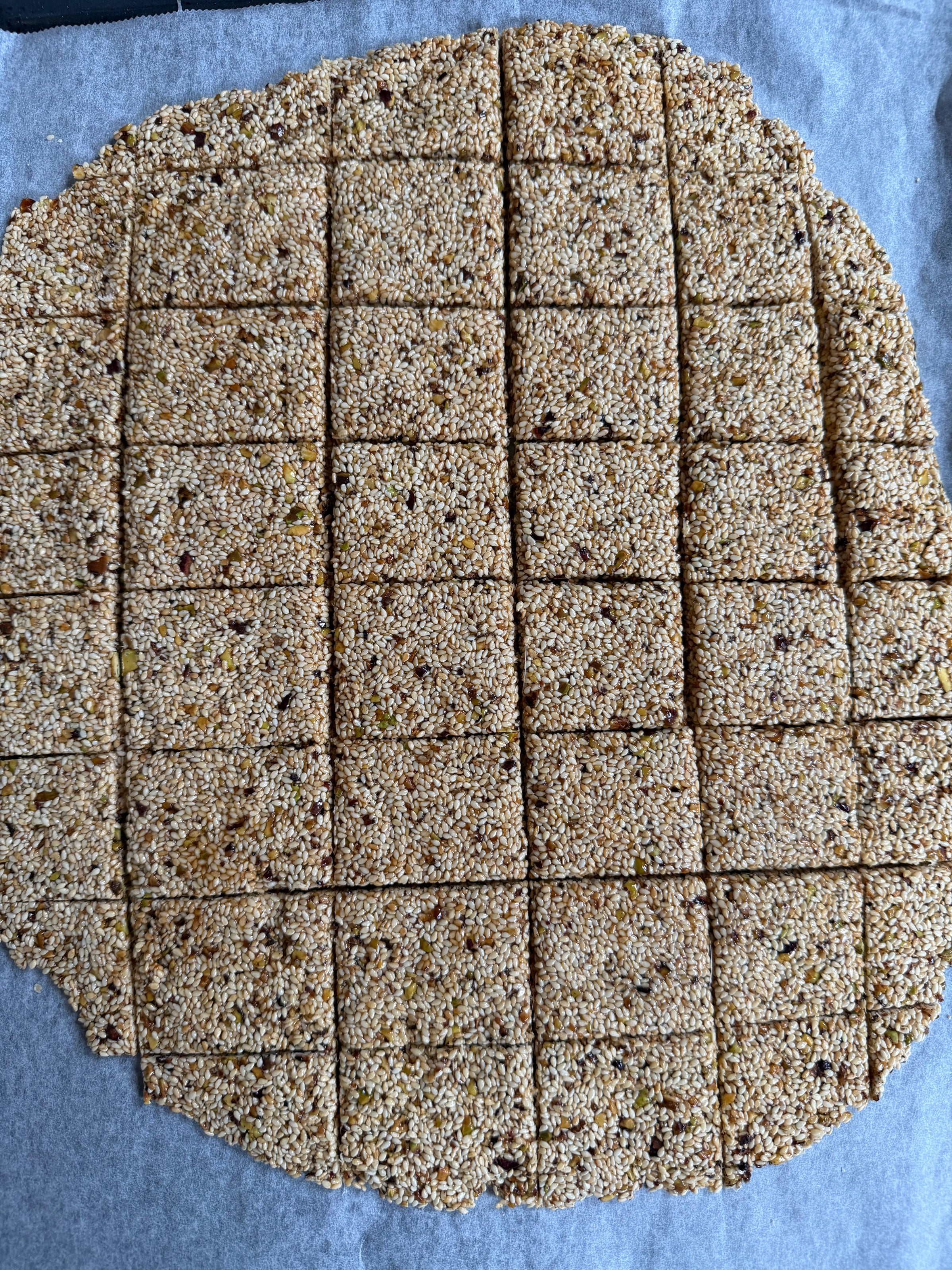 A large, round sheet of sesame seed brittle cut into square pieces, resting on parchment paper. The brittle has a golden color with visible seeds and nuts throughout.