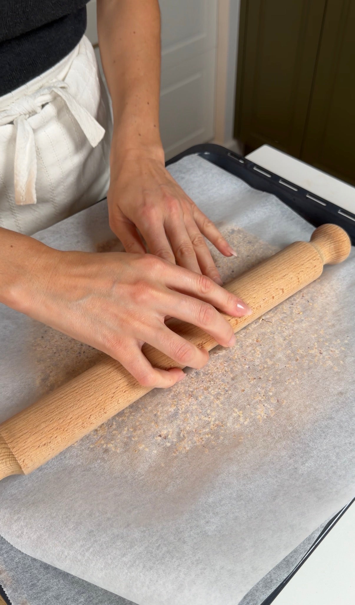 A person wearing a white apron uses a wooden rolling pin to flatten dough on parchment paper, with flour scattered on the surface.