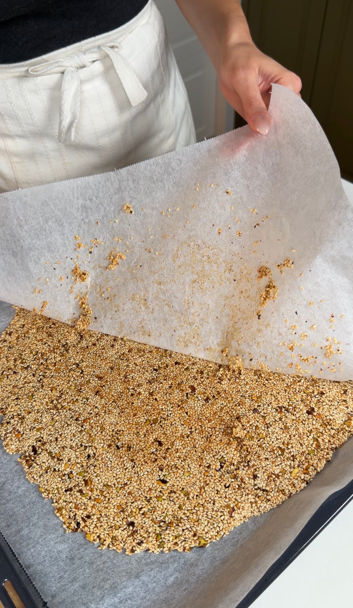 A person lifts parchment paper from a flat layer of sesame seed brittle on a baking tray, revealing the golden, crunchy surface underneath.