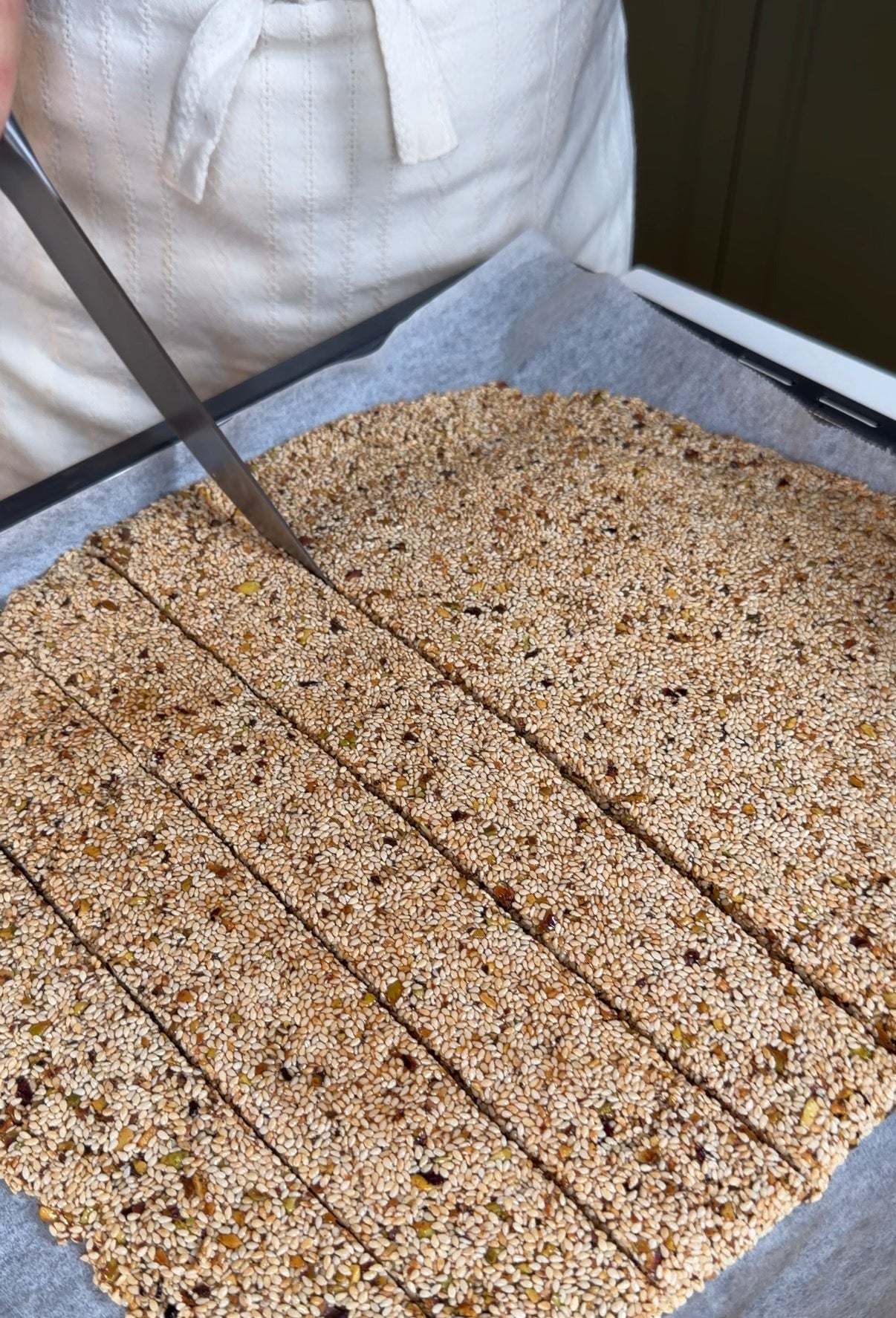 A person in a white apron uses a large knife to cut a baked sheet of sesame and seed brittle into rectangular strips on a parchment-lined baking tray.