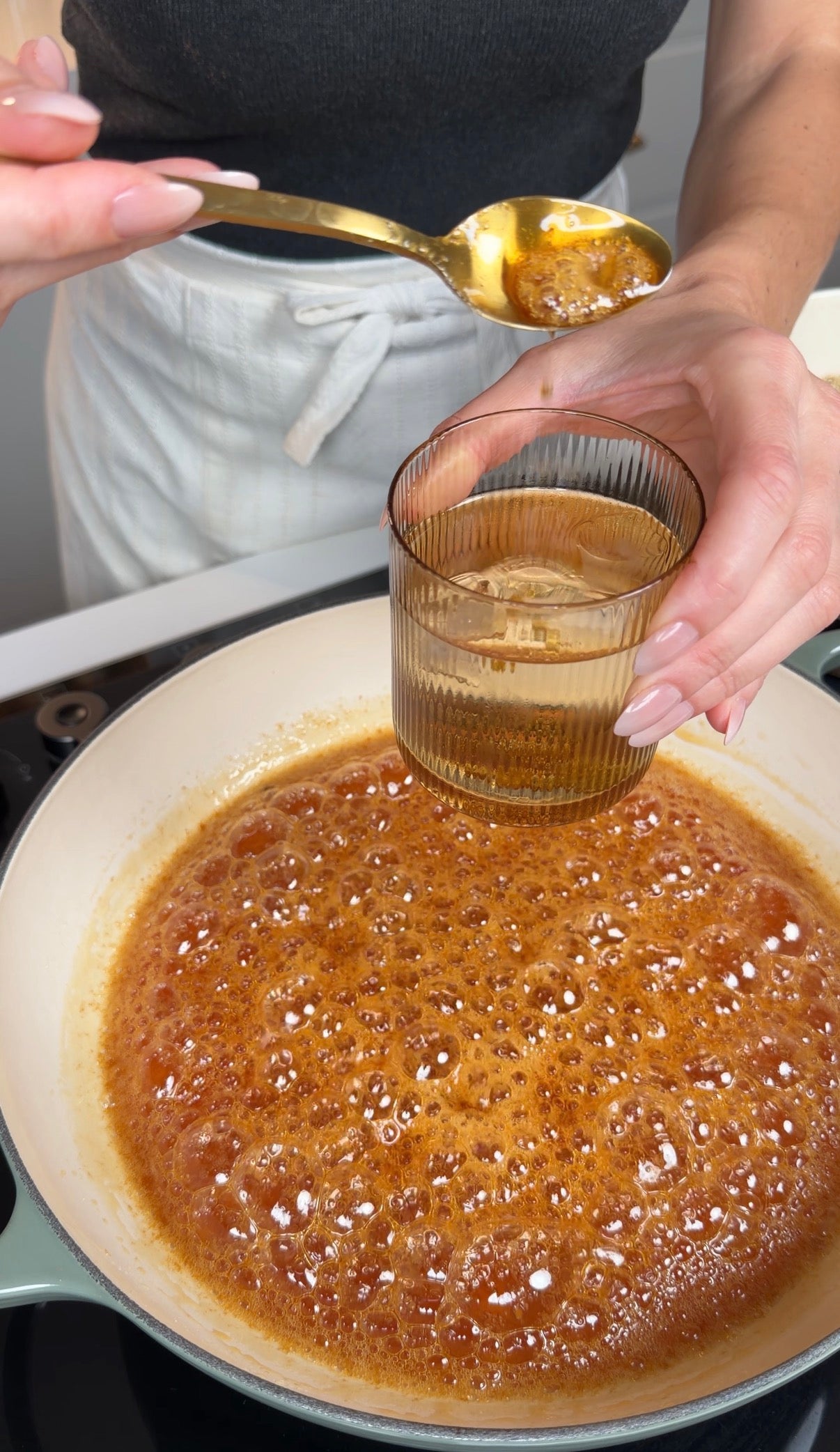 A person holds a glass of liquid and a spoonful of brown paste over a bubbling caramel mixture in a pan on the stove.