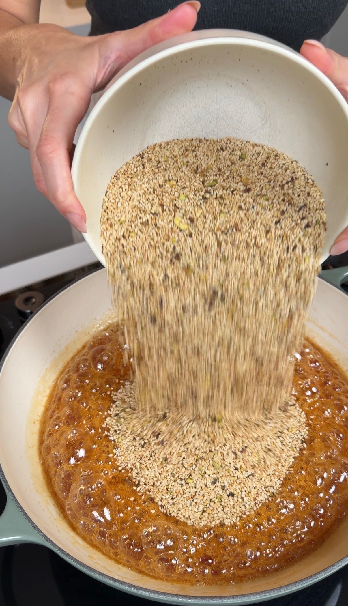 A person pours a bowl of mixed seeds into a pan filled with bubbling caramel on a stovetop, preparing to make a sesame seed brittle or similar treat.