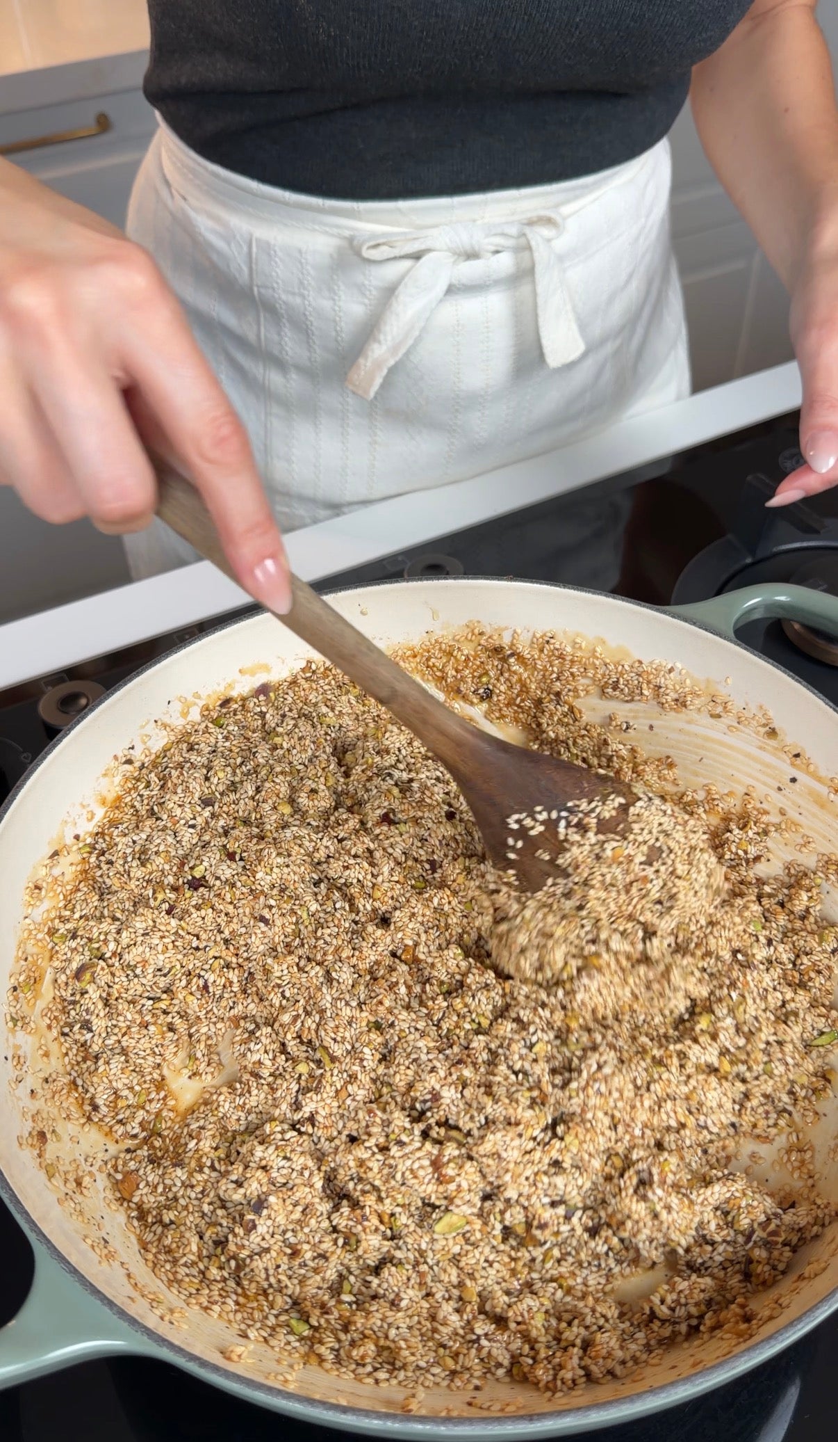 A person stirs a mixture of seeds and nuts in a large pan with a wooden spoon. They are wearing a gray shirt and a white apron, standing at a stove in a bright kitchen.