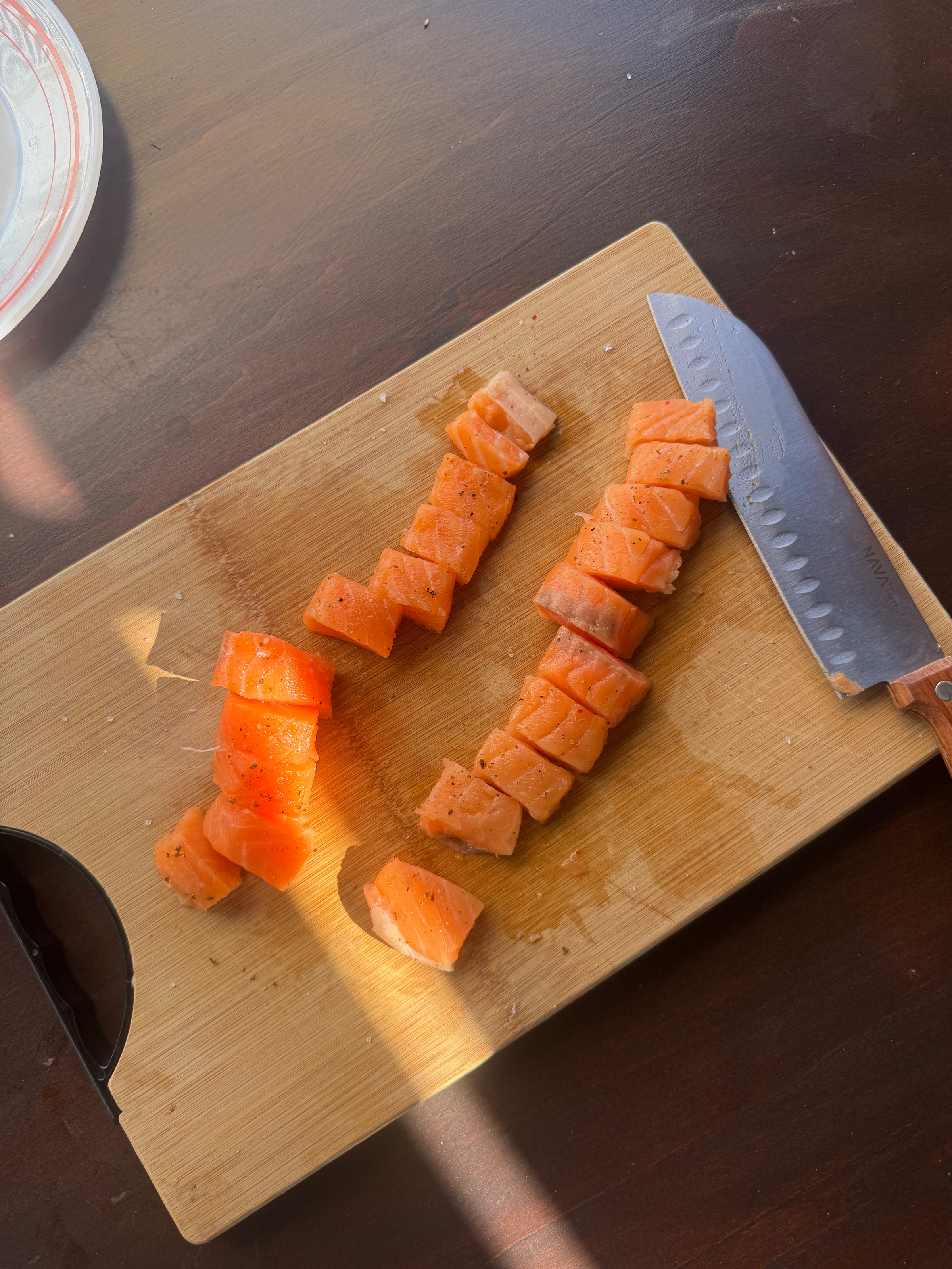 Cubed pieces of raw salmon are arranged on a wooden cutting board next to a large kitchen knife with a wooden handle. A plate is partially visible in the upper left corner. Sunlight shines across the board.