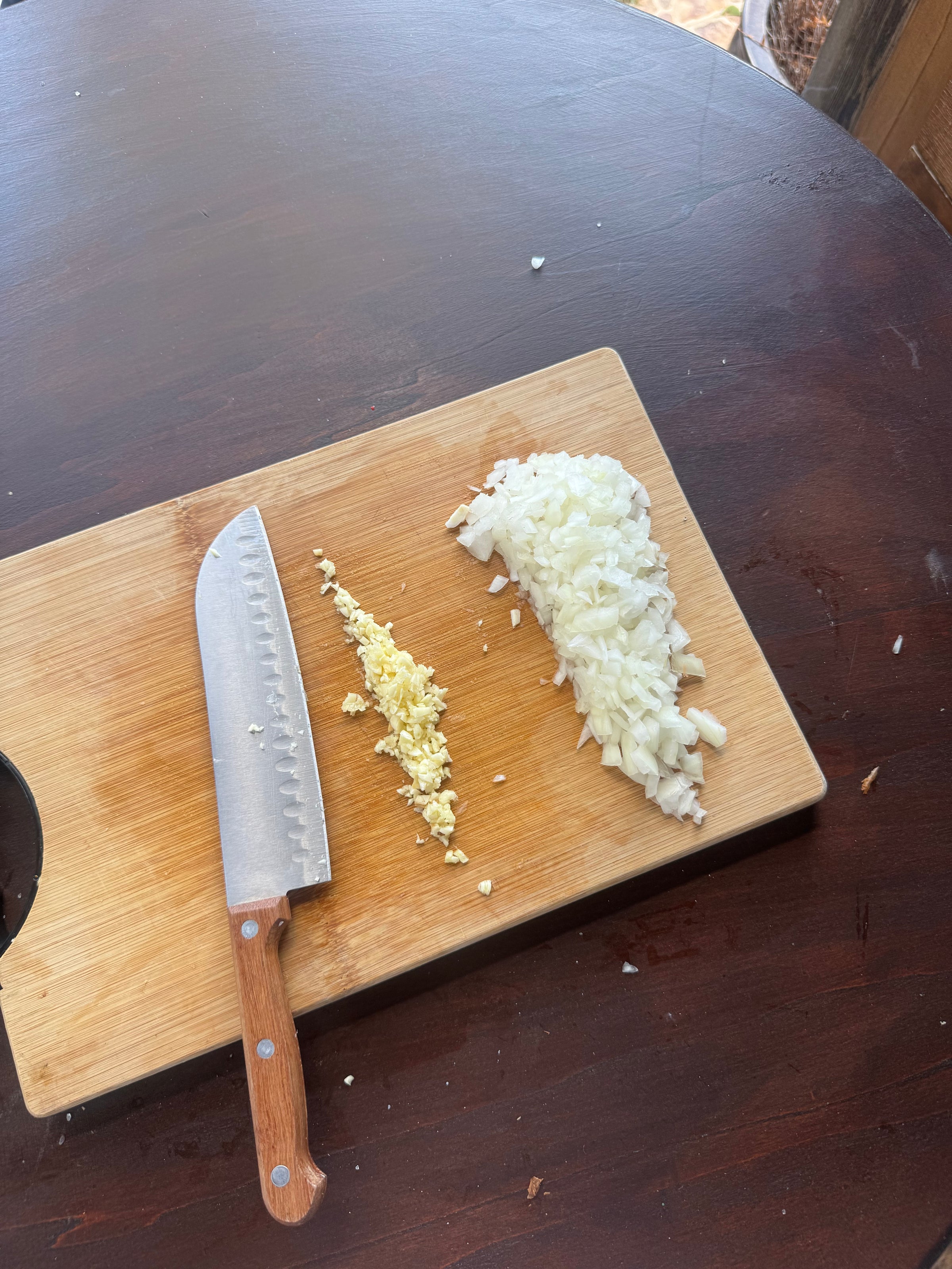 A wooden cutting board on a dark table holds a knife, a pile of minced garlic, and a pile of chopped onions. Some onion pieces are scattered on the board.
