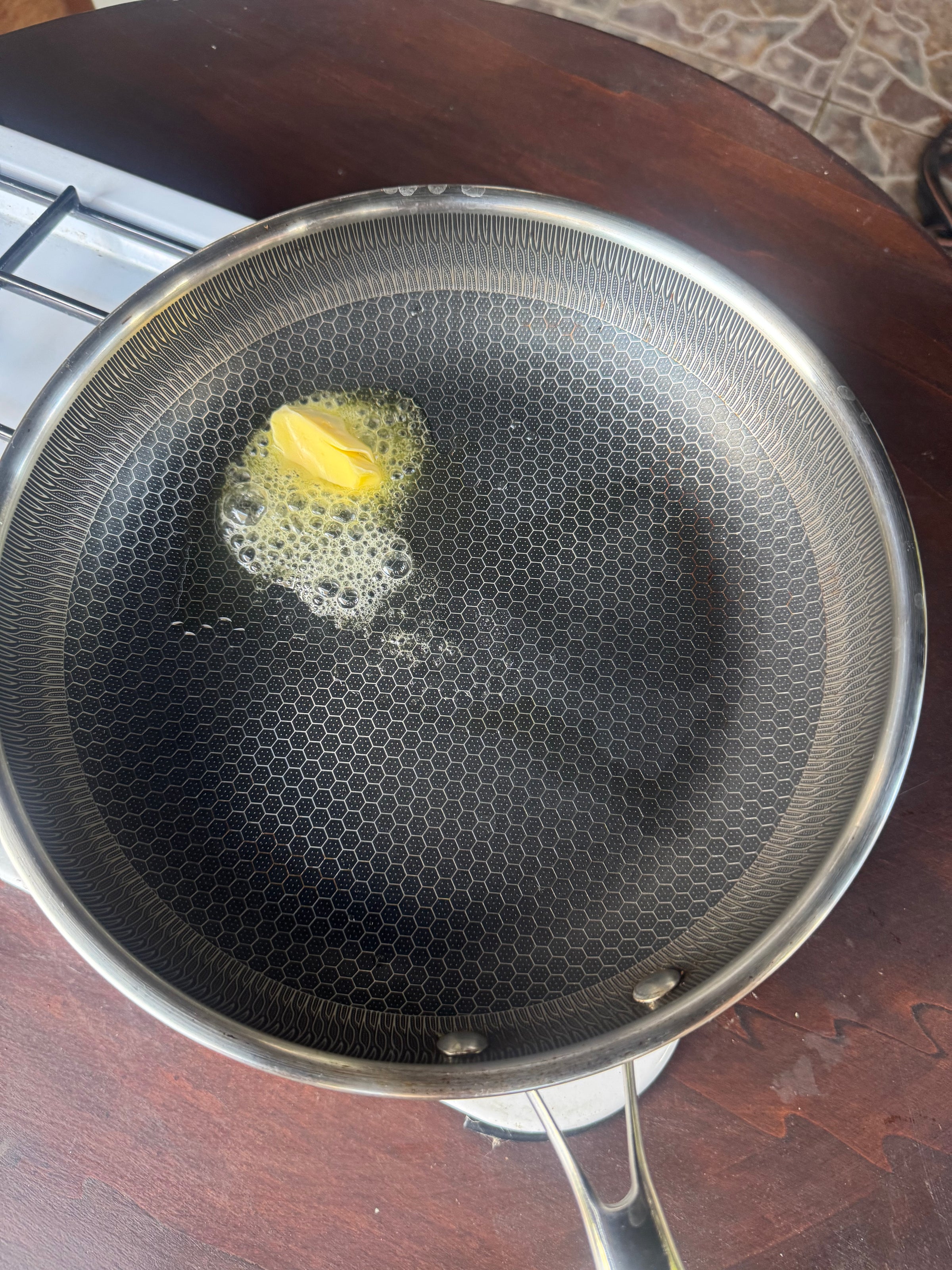 A piece of butter melting in a honeycomb-textured frying pan on a stove, with bubbles forming around the butter. The pan sits on a dark wooden surface.