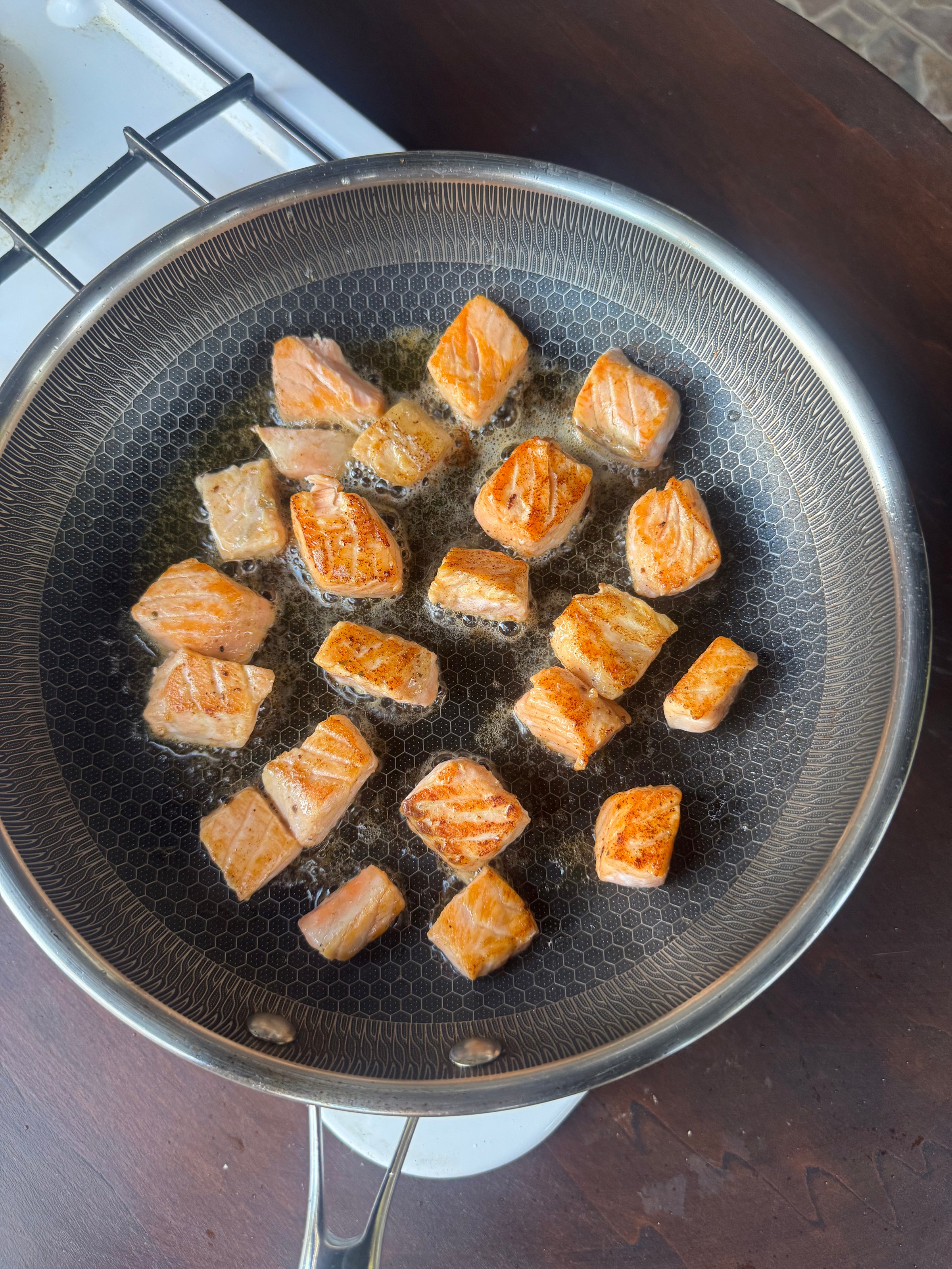 Cubed pieces of salmon are being pan-fried in oil on a stovetop. The salmon is golden brown and sizzling in a round, nonstick frying pan.