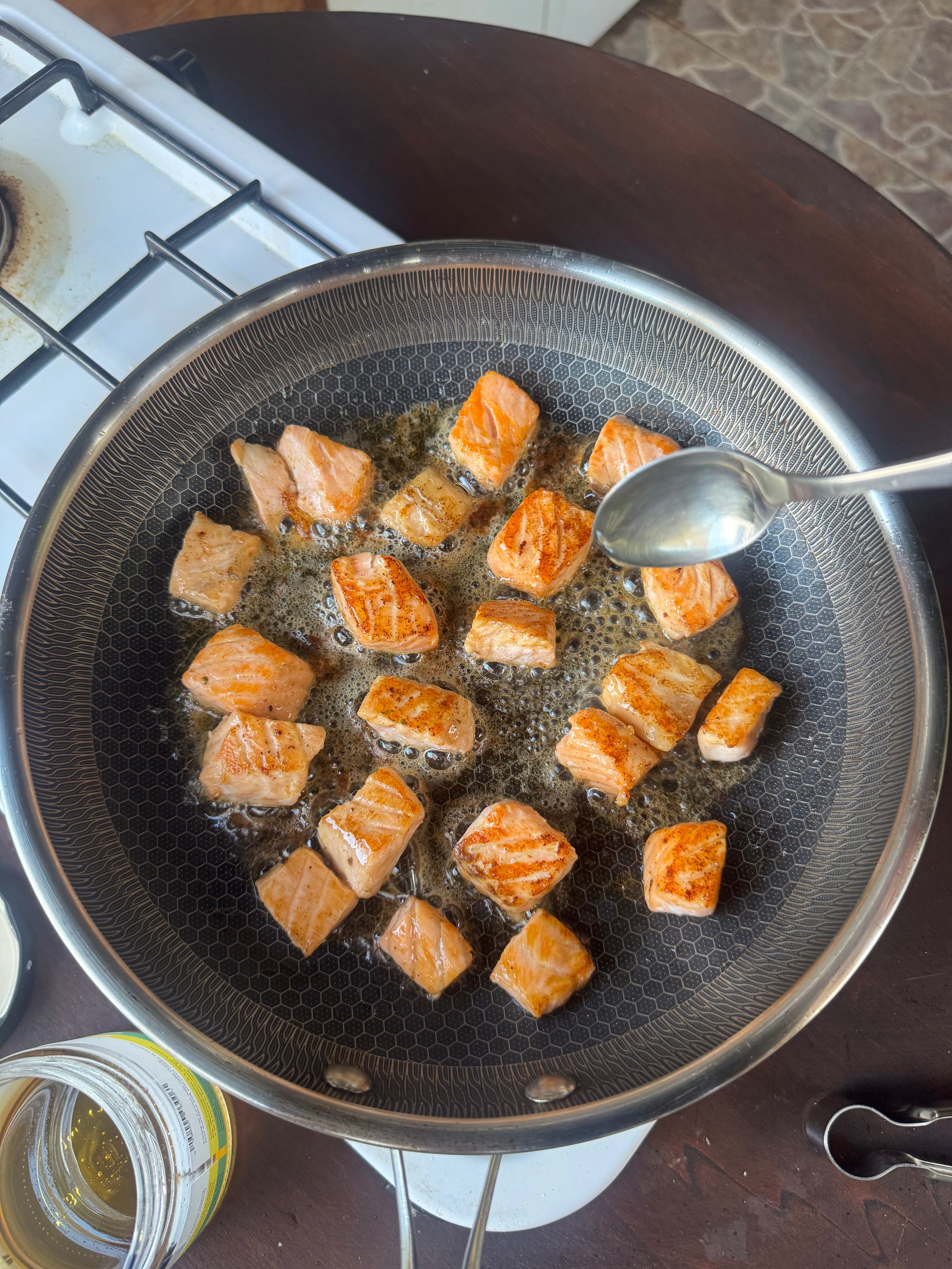 Cubed salmon pieces are being pan-fried in oil in a frying pan on a stove. A hand is holding a spoon above the pan, possibly to baste the salmon or scoop something. A jar and stove burner are visible nearby.