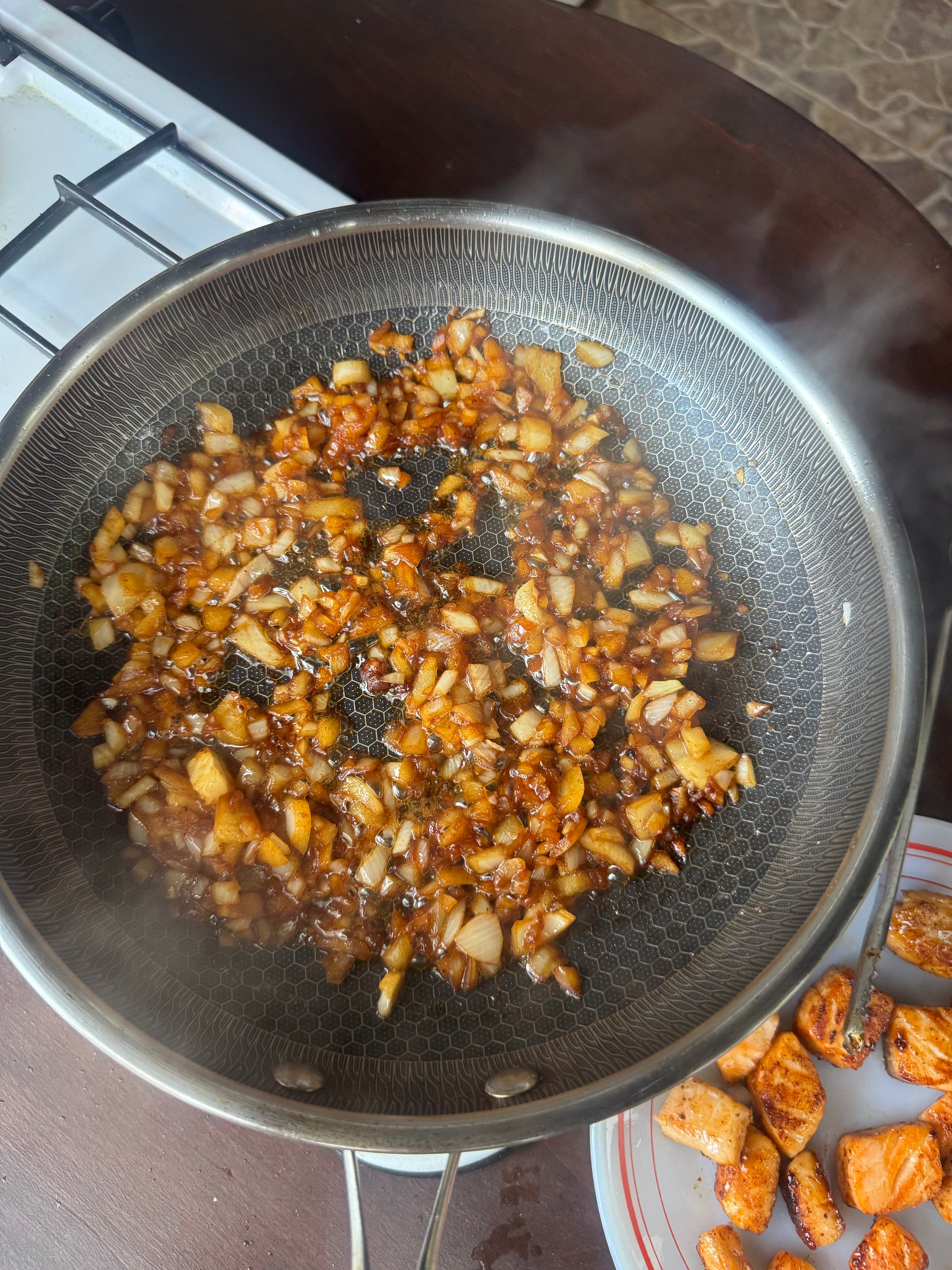 Chopped onions are being sautéed in a frying pan with spices. The mixture is browning, and some cooked pieces of chicken are visible on a plate nearby. Steam rises from the pan.