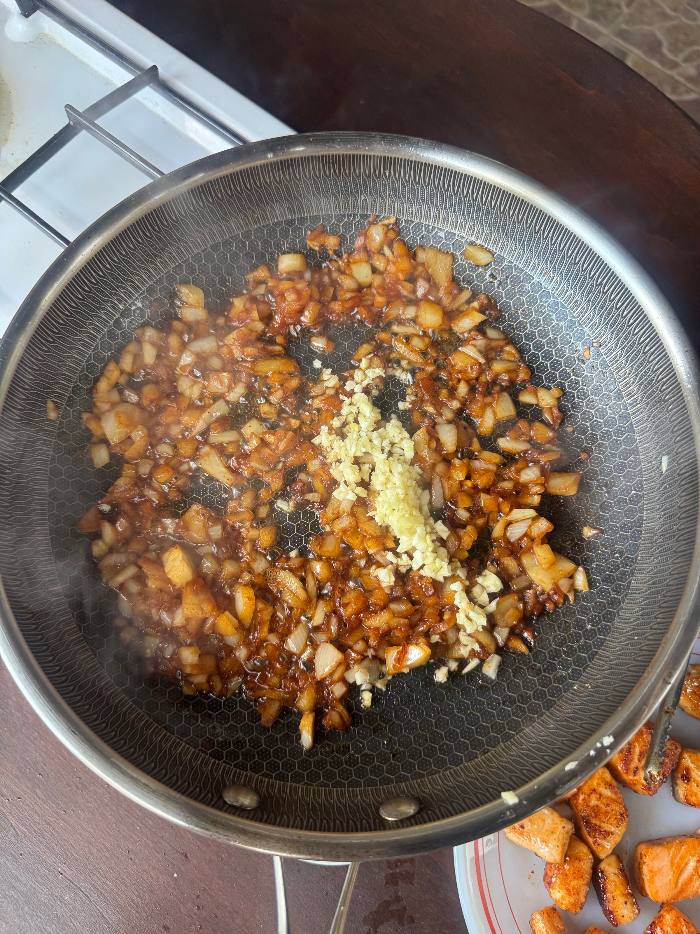 Chopped onions sautéing in a frying pan with a pile of minced garlic added in the center. Theres some chopped food on a plate nearby. Steam rises from the hot mixture.