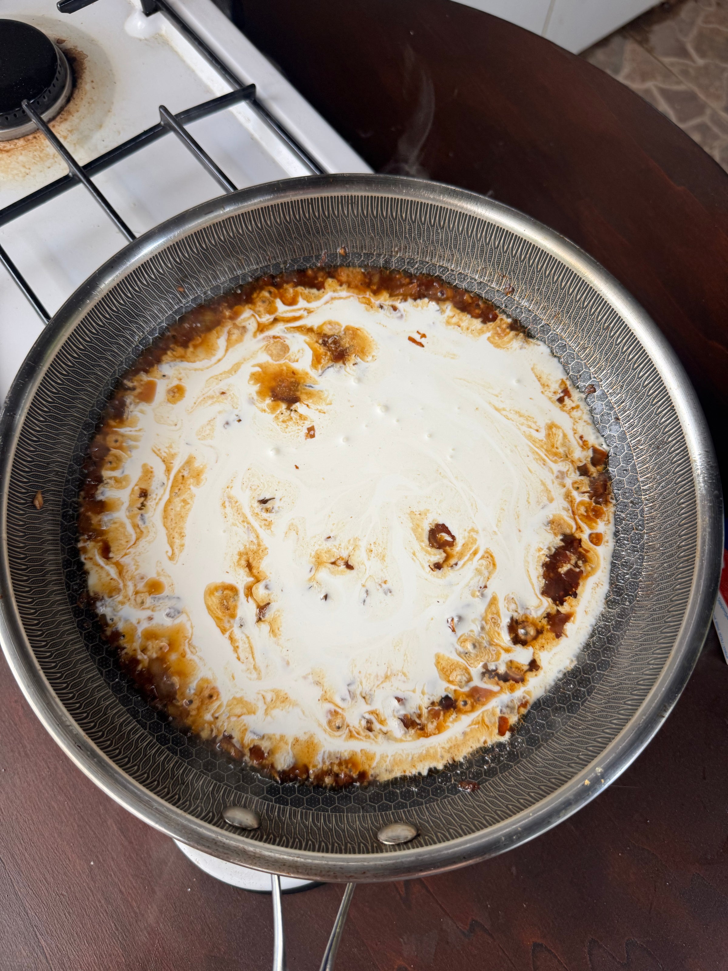 A metal pan on a stove contains cream and brown sauce swirling together, likely being heated and mixed for cooking. The stove and part of a dark wooden surface are visible in the background.