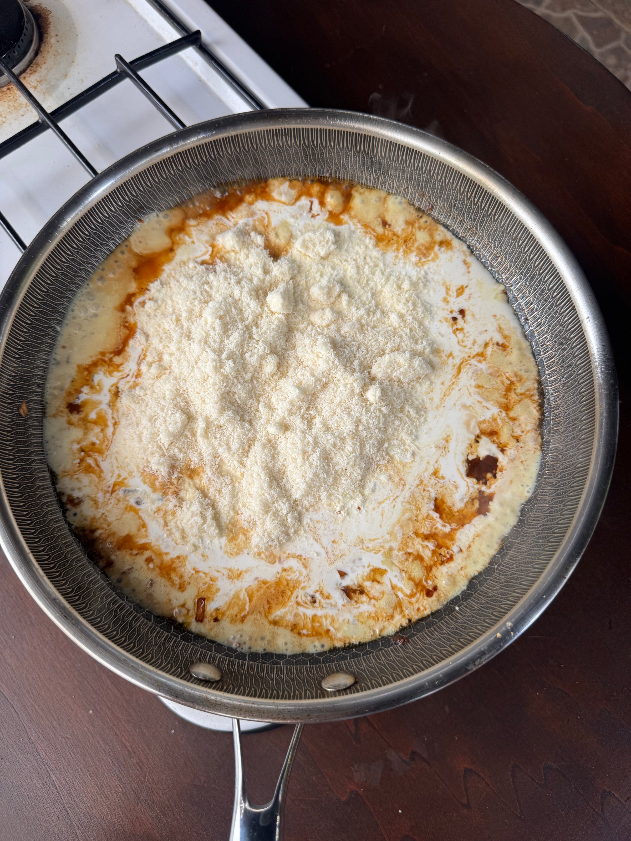 A frying pan on a stove containing cream, grated cheese, and some spices, starting to melt and mix together for a sauce.