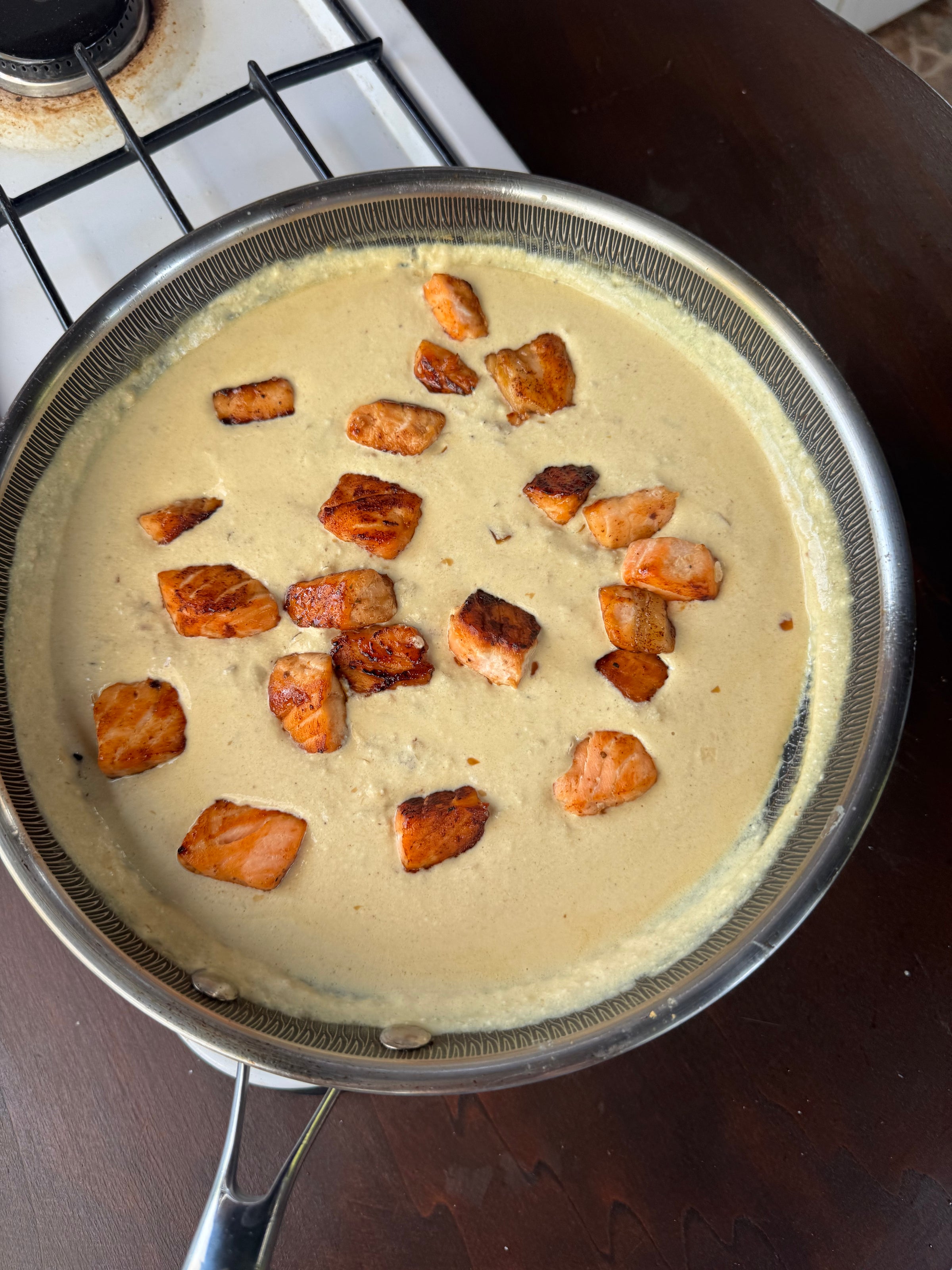 Chunks of seared salmon in a creamy white sauce, cooking in a frying pan on a stovetop next to a dark wooden counter.