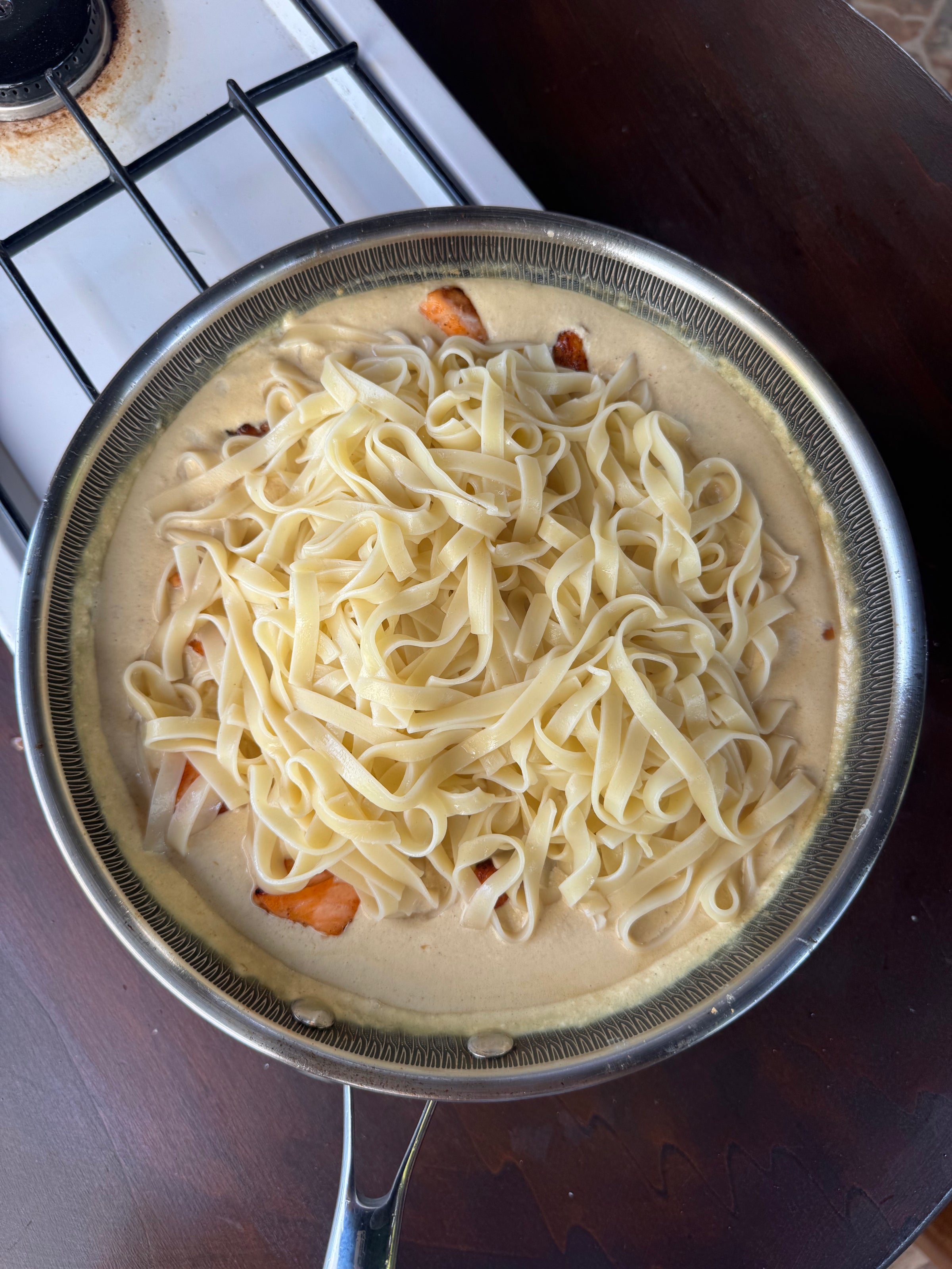 A pan with creamy sauce, pieces of cooked chicken, and a pile of uncooked fettuccine pasta sits on a stove, ready to be mixed together.