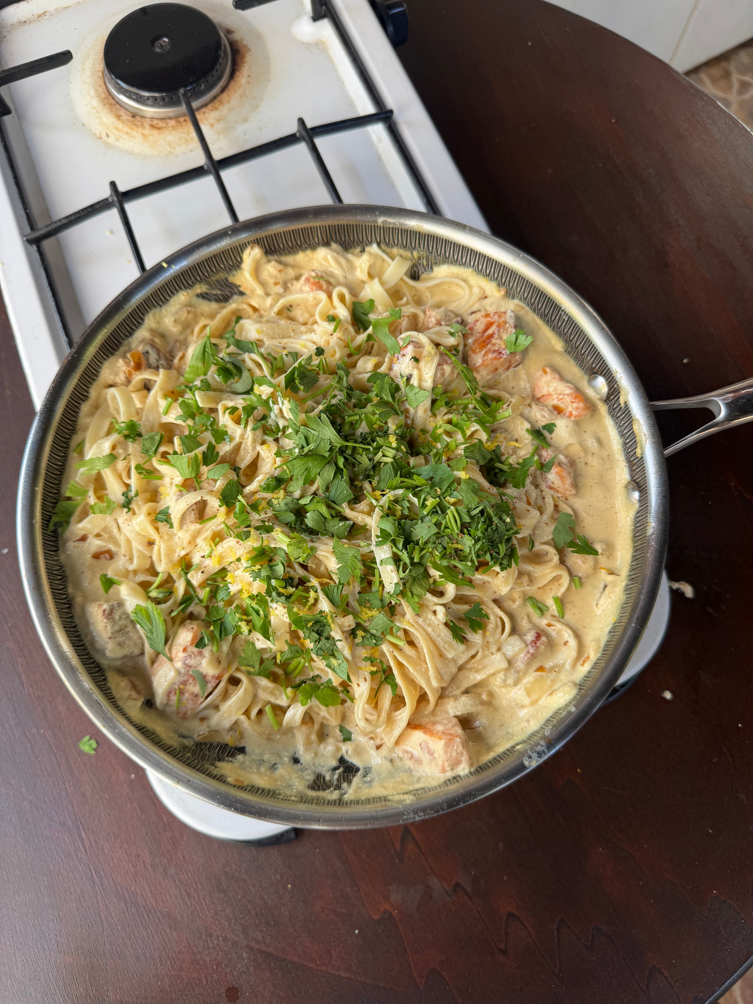 A skillet of creamy pasta with pieces of chicken and chopped fresh herbs sits on a dark wooden table next to a white gas stove.