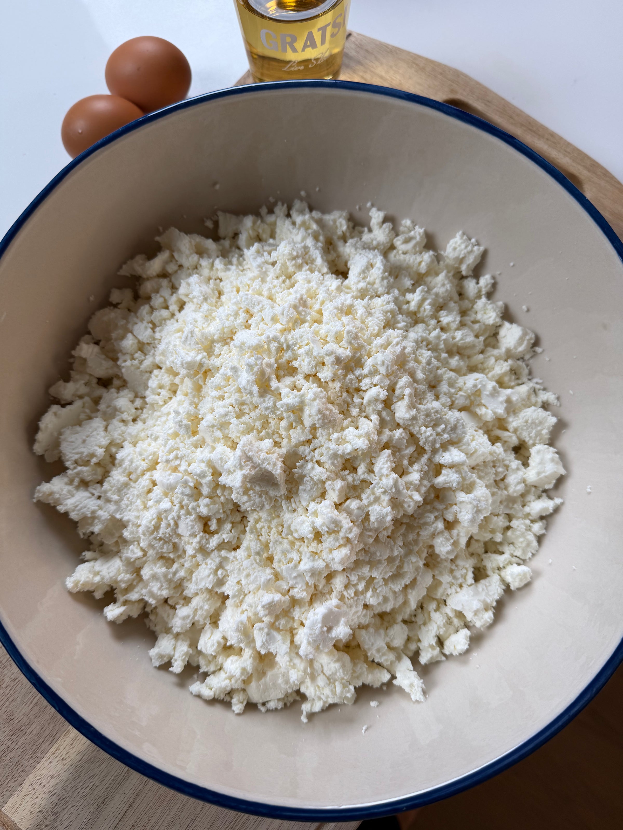 A large bowl filled with crumbled white cheese sits on a wooden surface. Nearby, there are two brown eggs and a bottle labeled GRATS.