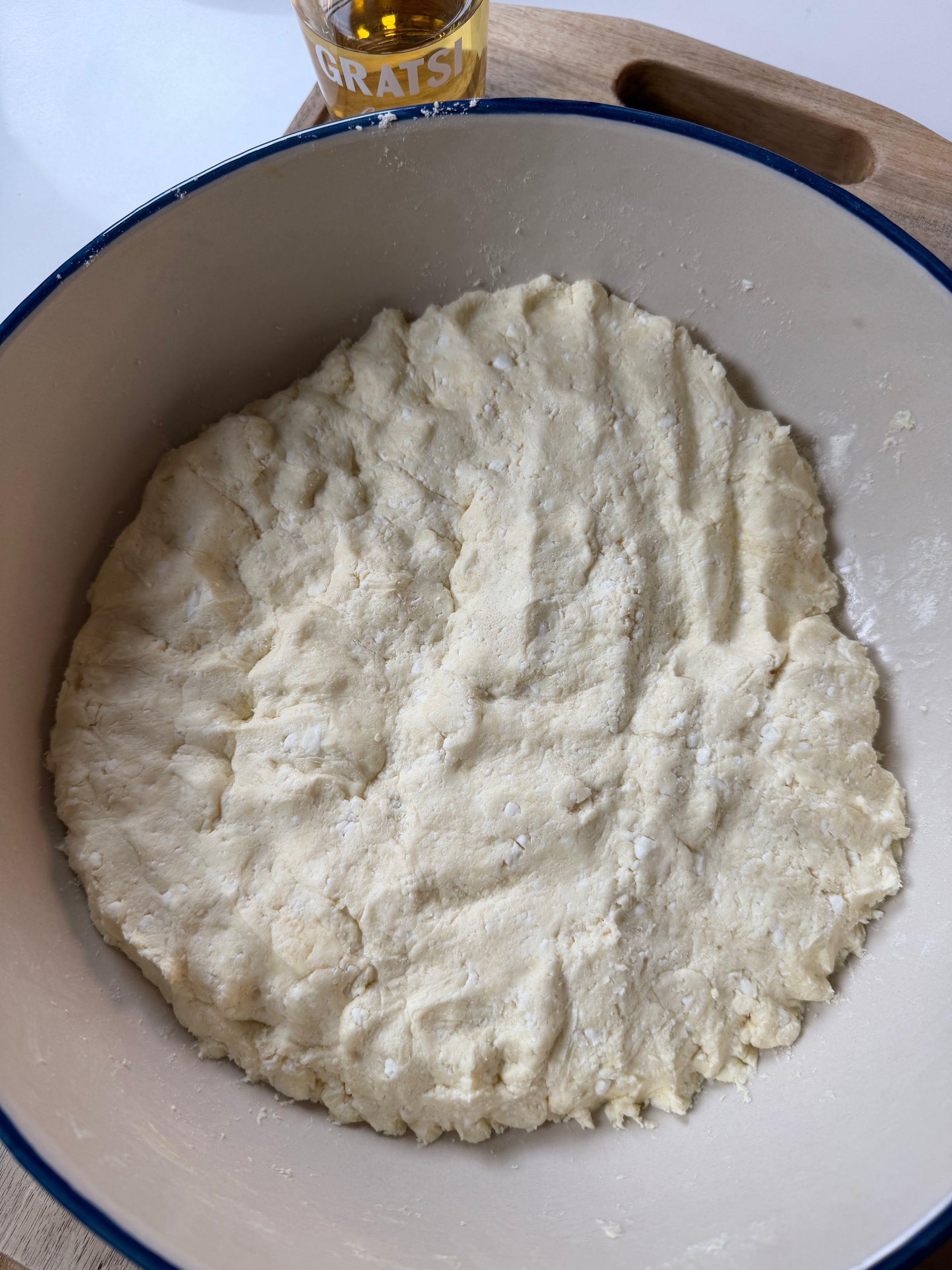 A bowl of unbaked dough pressed flat, resting on a wooden cutting board. The edge of a glass with the word GRATSI is partially visible at the top of the image.