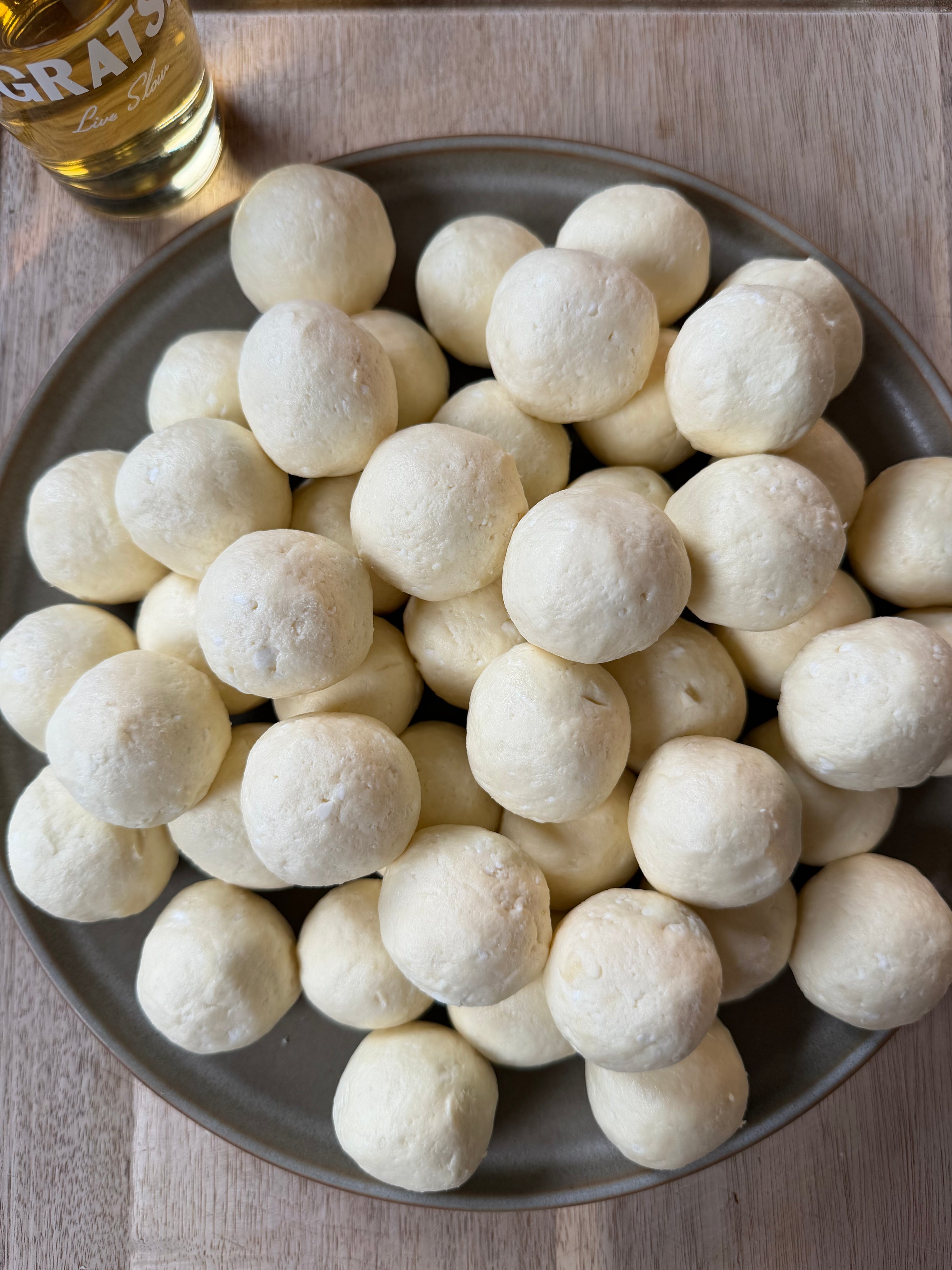 A plate filled with round, golden-white cheese bread rolls sits on a wooden surface, with part of a glass bottle visible in the upper left corner.