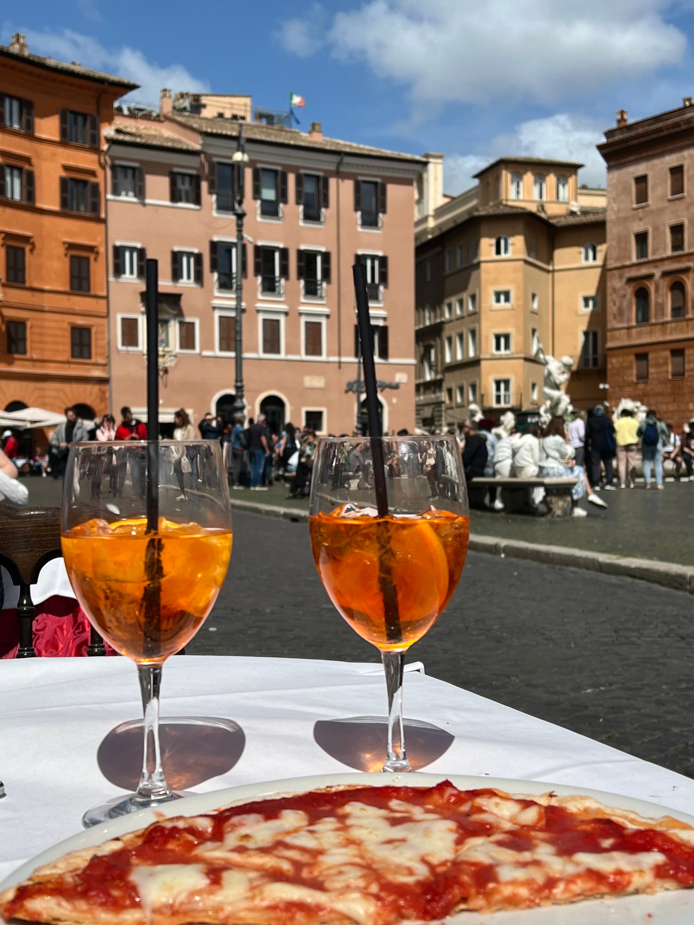Two glasses of orange Aperol spritz and a margherita pizza on a table outdoors, with people, historic buildings, and a fountain in a sunny Italian plaza in the background.