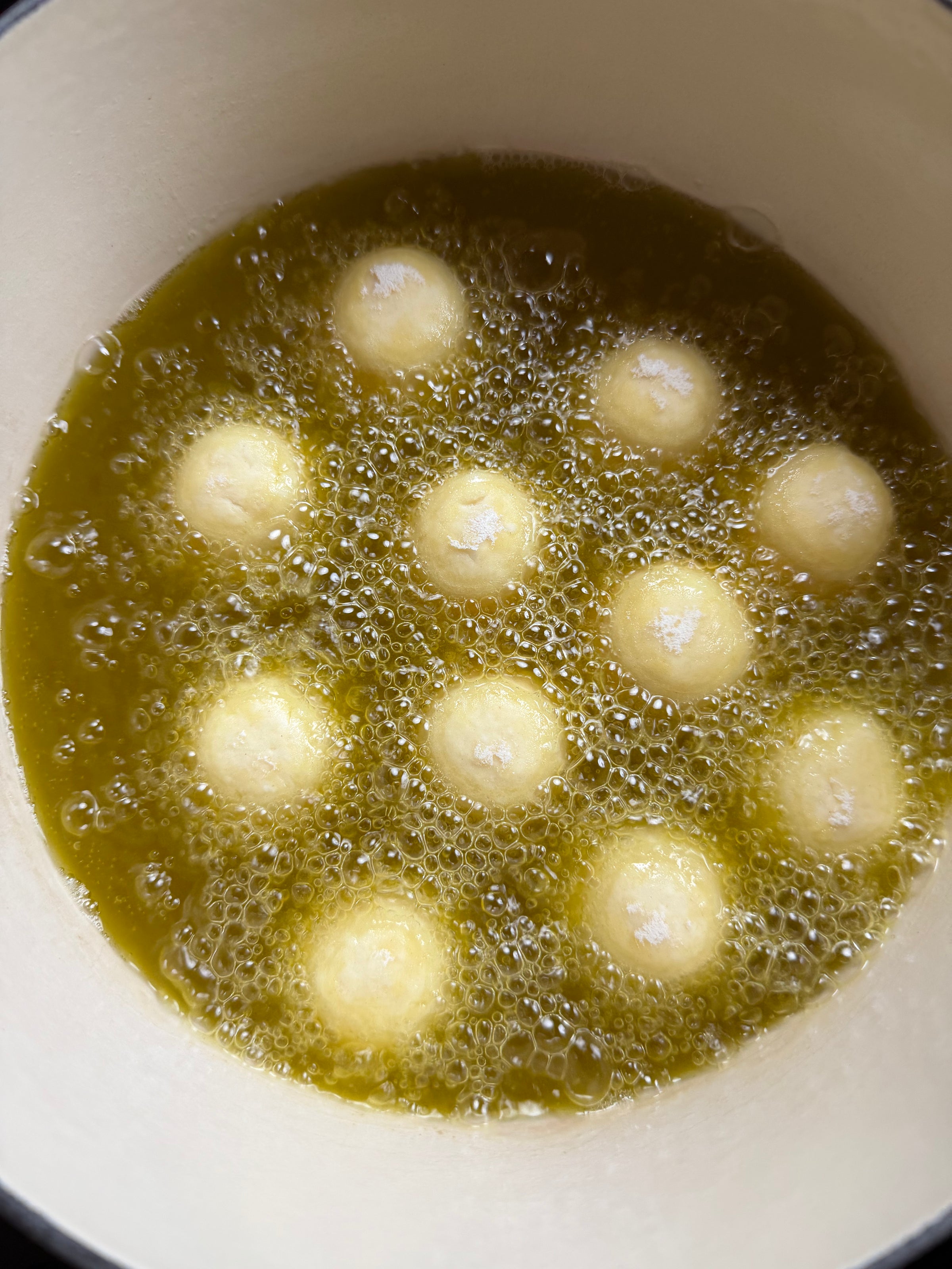 Round dough balls frying in bubbling hot oil, viewed from above in a white pot.