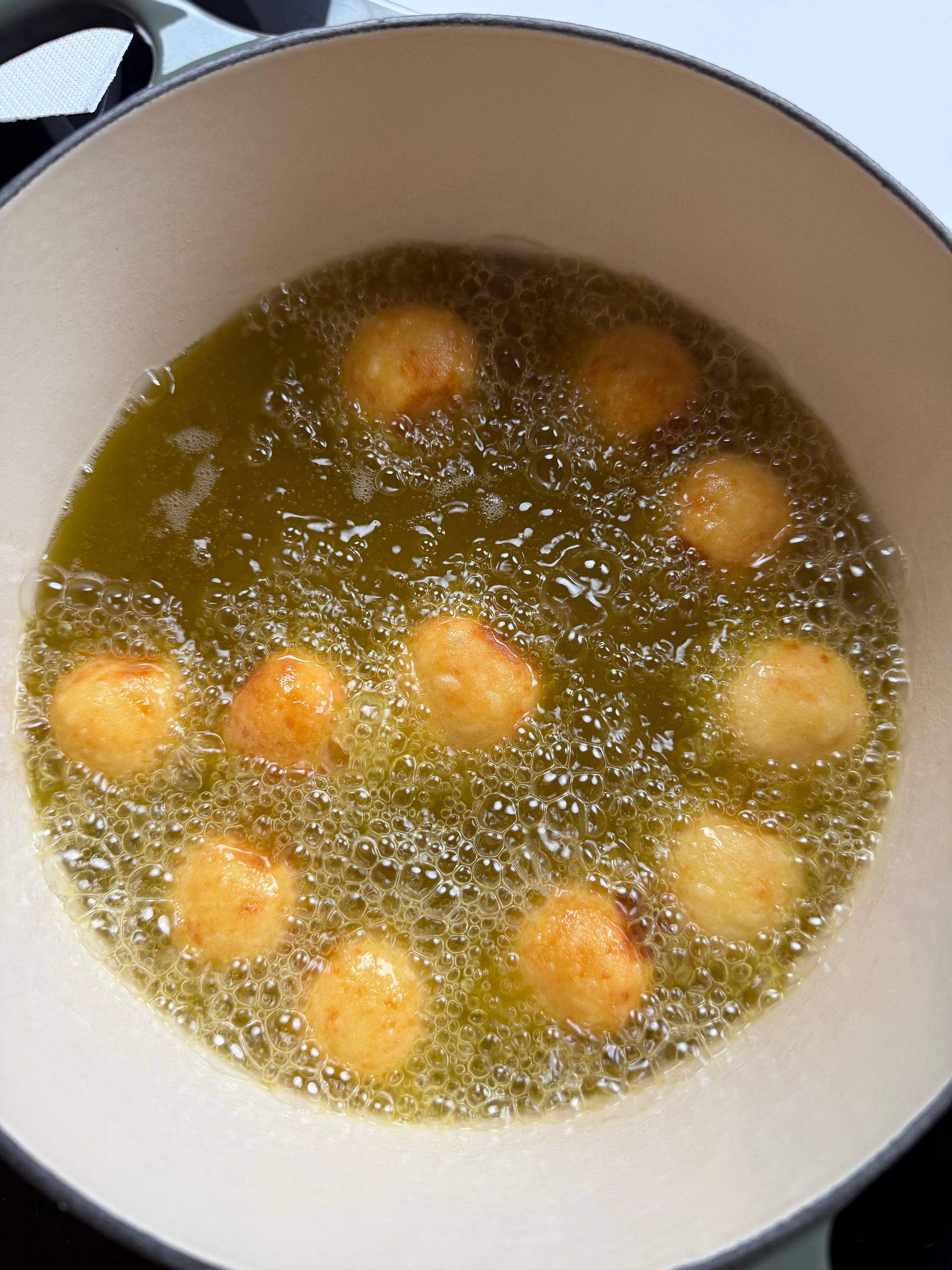 Round dough balls are being deep-fried in hot oil inside a white pot. The dough balls are turning golden brown as they cook, with bubbles forming around them.