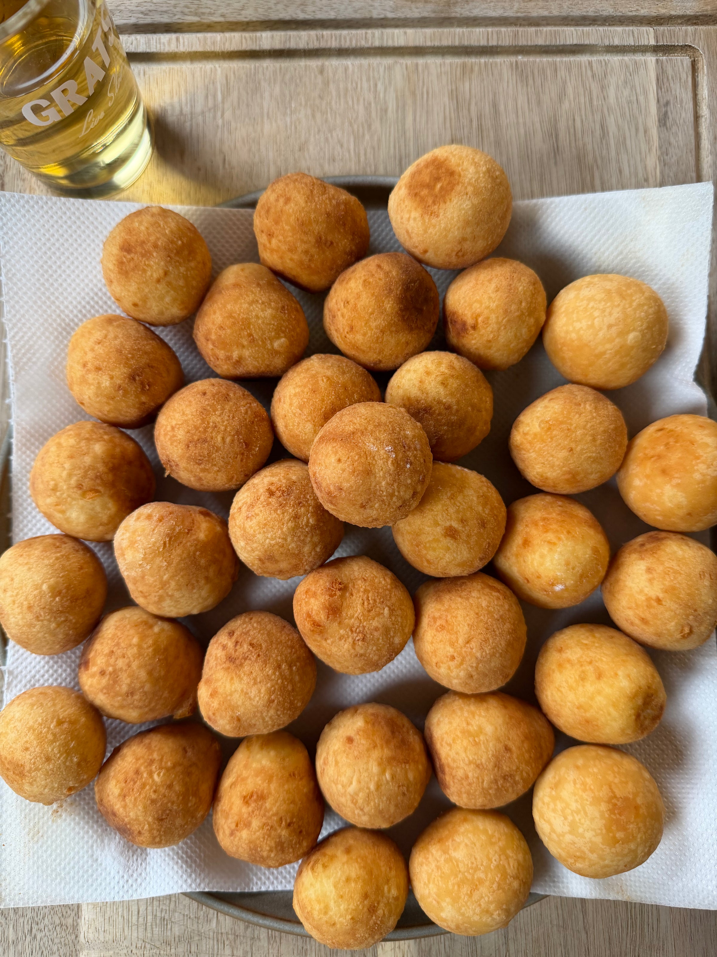 A plate filled with golden-brown, round fried balls sits on a paper towel atop a wooden surface; a partial glass of light-yellow liquid is visible in the top left corner.