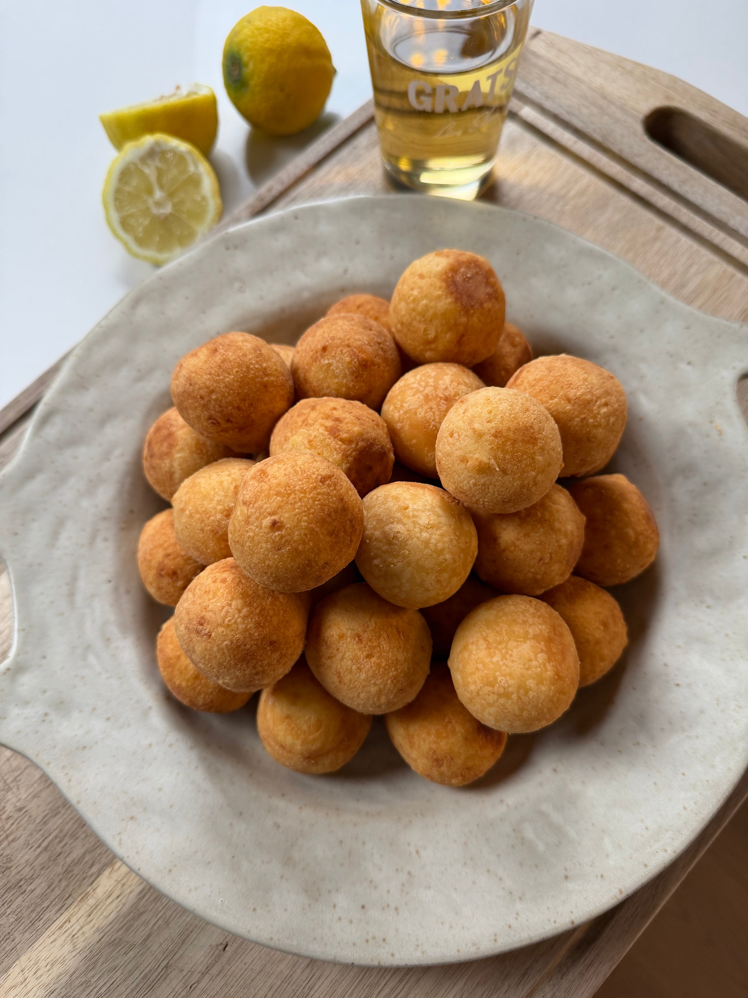 A plate piled with golden, round cheese balls sits on a wooden tray. In the background, there are lemon halves, a whole lemon, and a glass with a light-colored beverage.