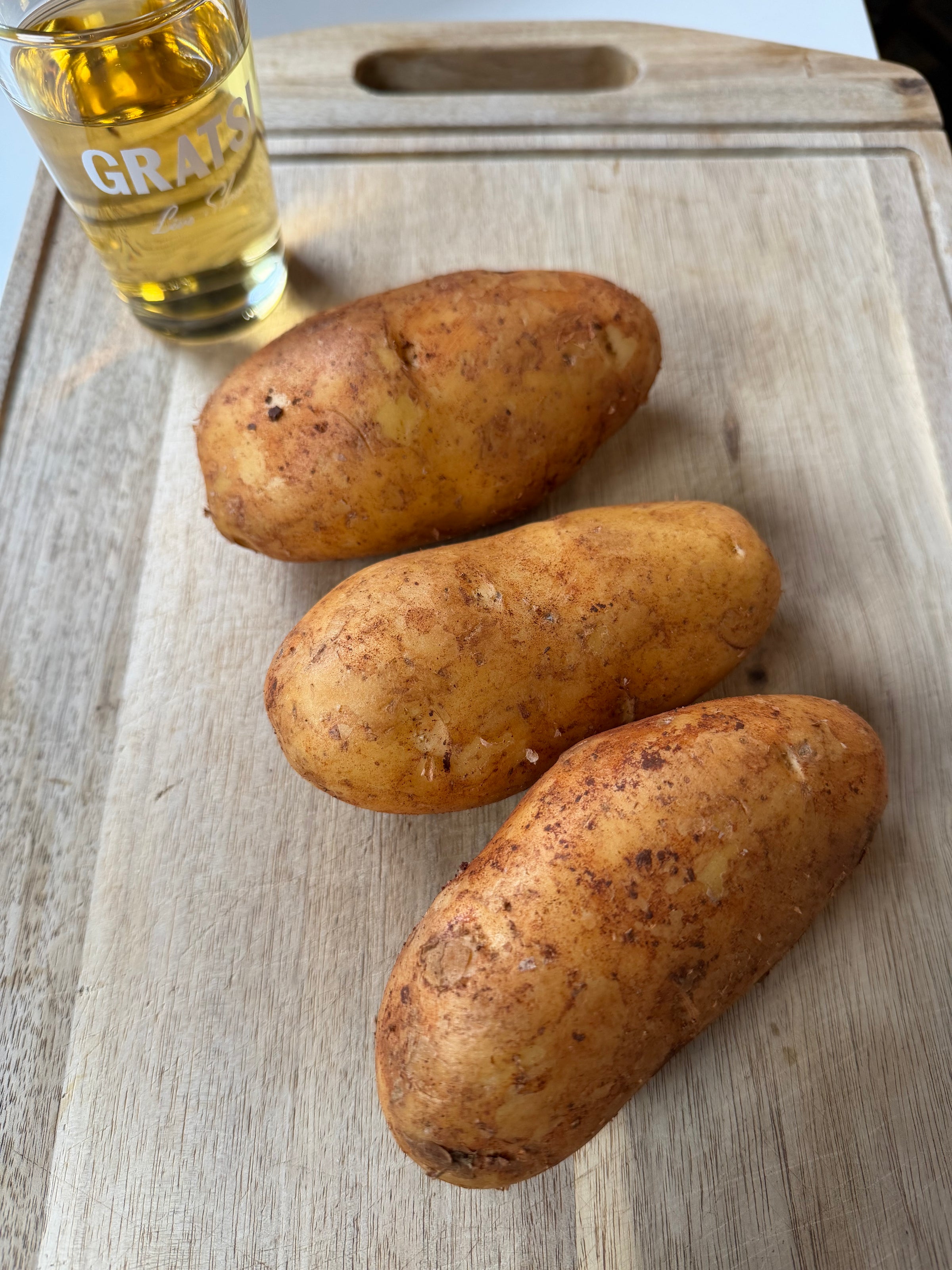 Three unpeeled potatoes are placed on a wooden cutting board. In the background, there is a clear glass of golden liquid labeled GRATI, partially visible.