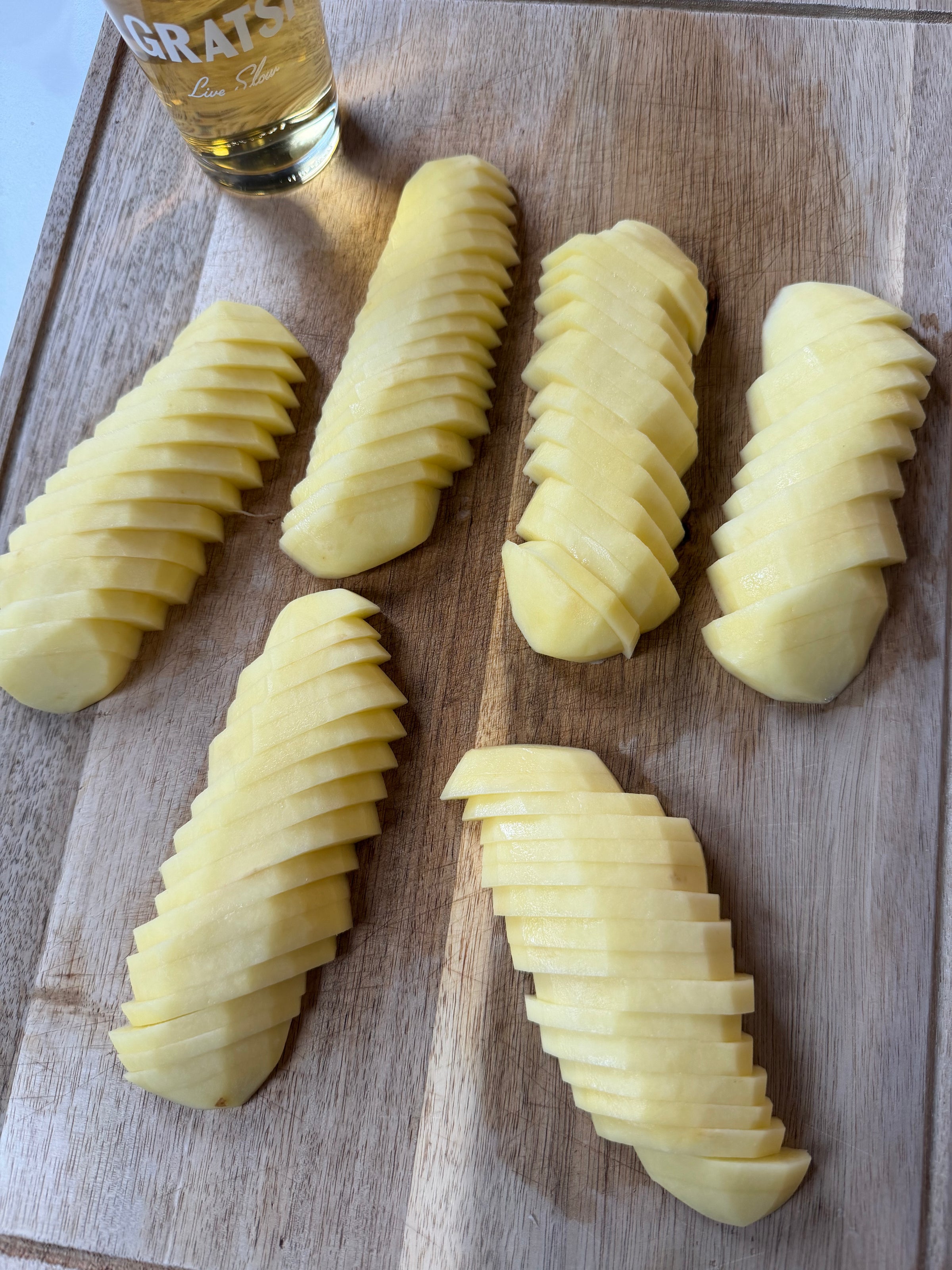 Six peeled potatoes are thinly sliced and neatly fanned out on a wooden cutting board. A glass of light-colored beverage, partially visible, is placed in the top left corner.