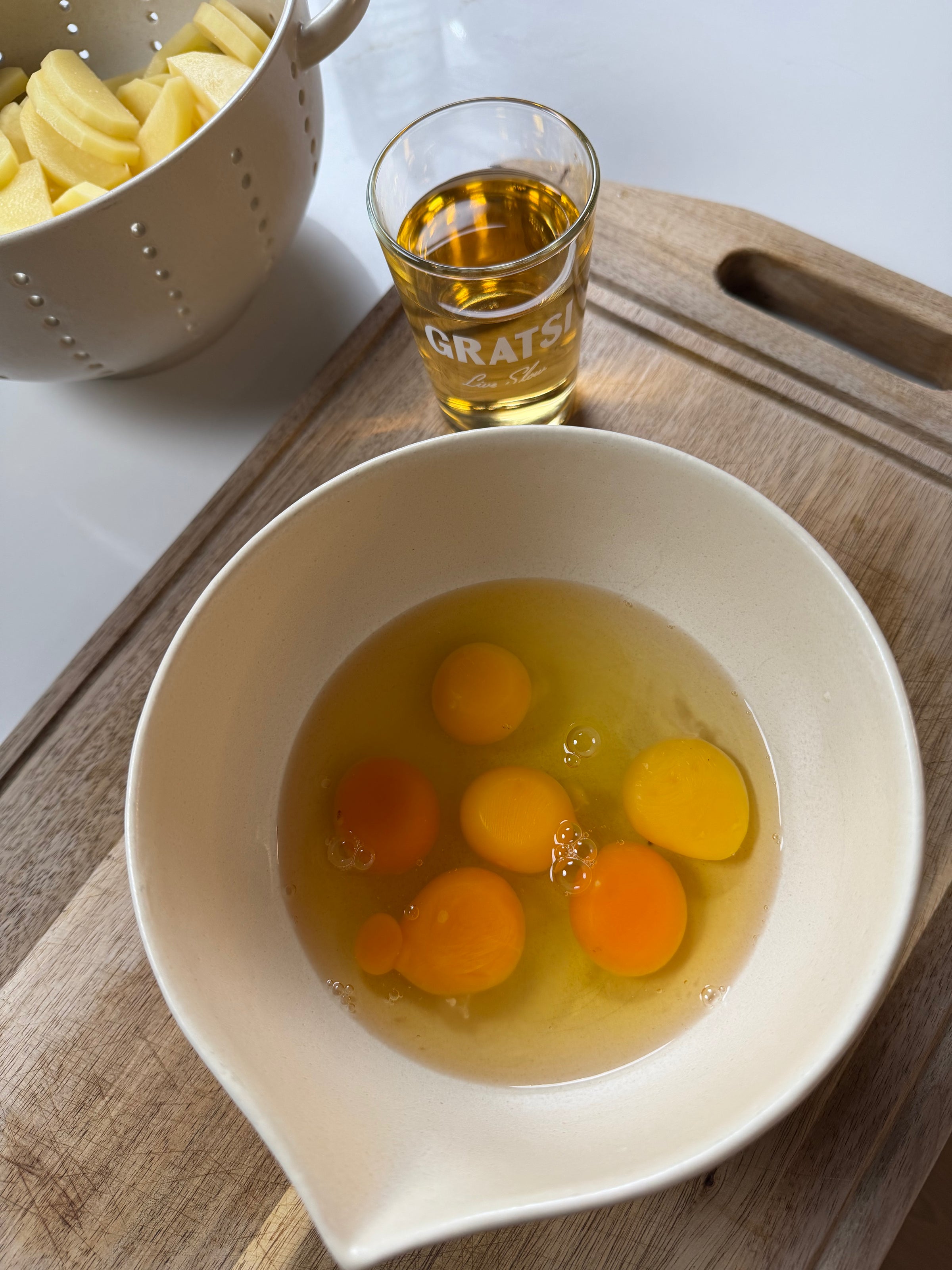 A white bowl with six cracked eggs sits on a wooden cutting board beside a shot glass of amber liquid labeled GRATS! and a colander of sliced apples.