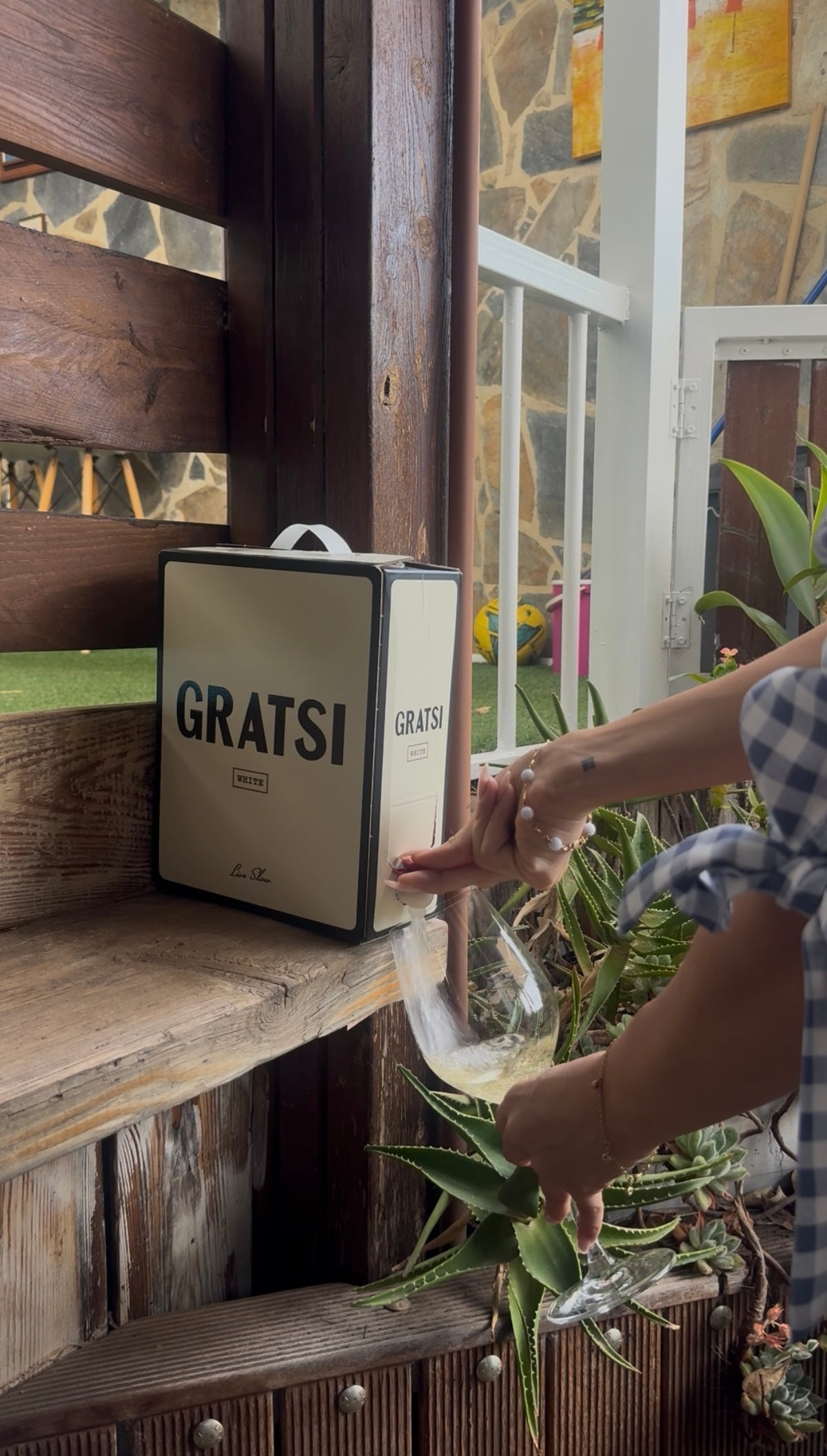 A person in a checkered shirt pours liquid from a GRATS! box into a glass, outdoors next to plants and wooden seating.