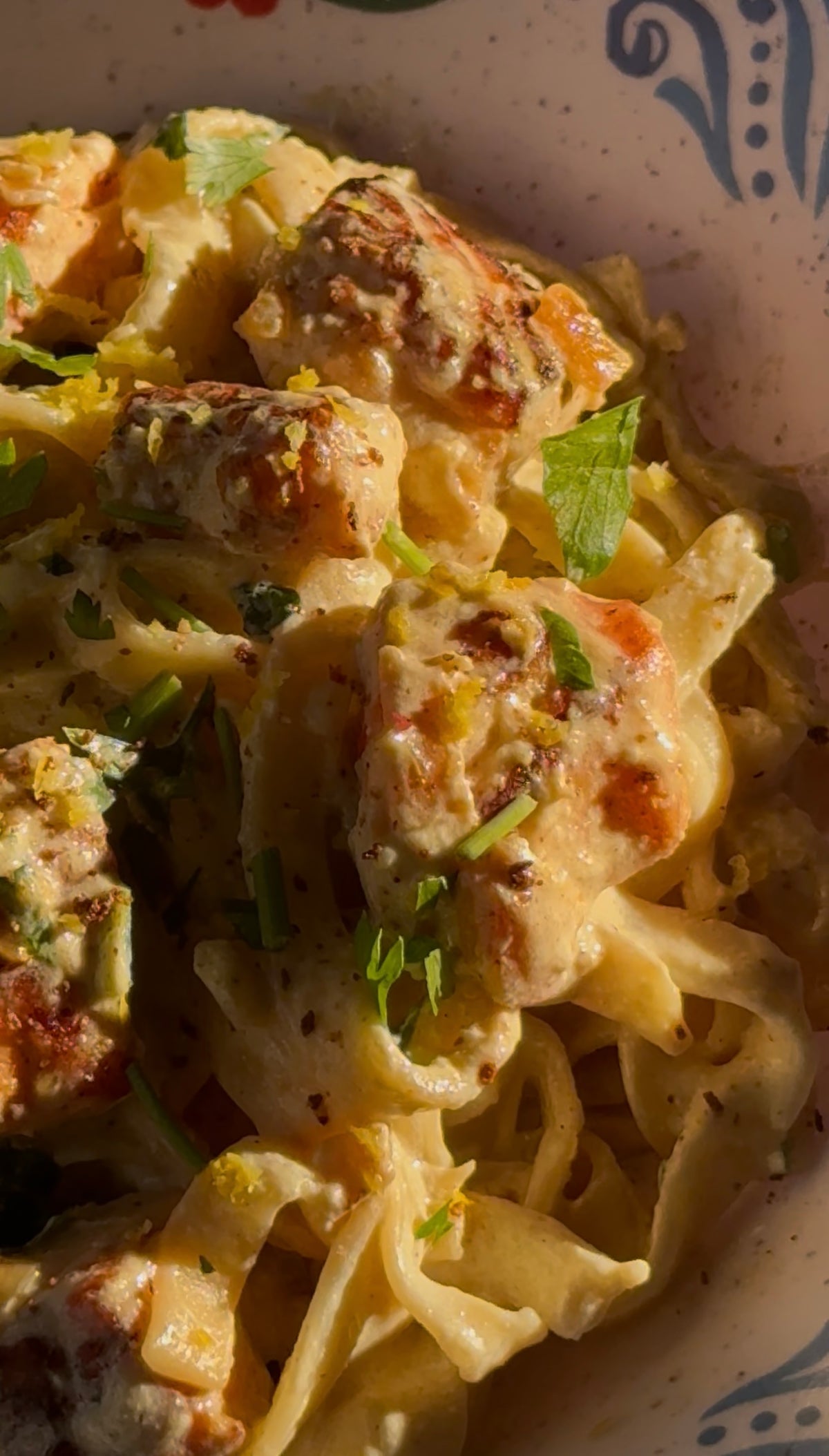 Close-up of creamy pasta topped with grilled chicken pieces, chopped herbs, and lemon zest, all bathed in warm sunlight.