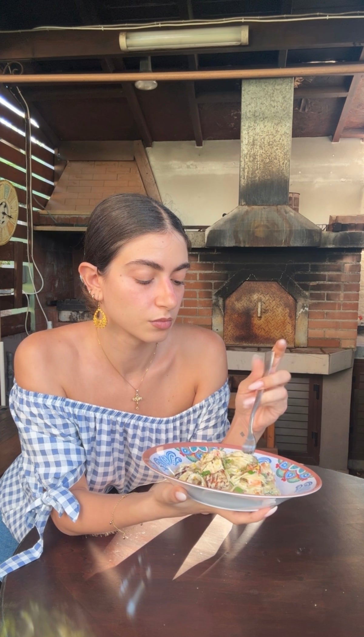 A woman in a blue and white off-shoulder top eats food from a colorful bowl, sitting at a wooden table in an outdoor kitchen with a brick oven behind her.