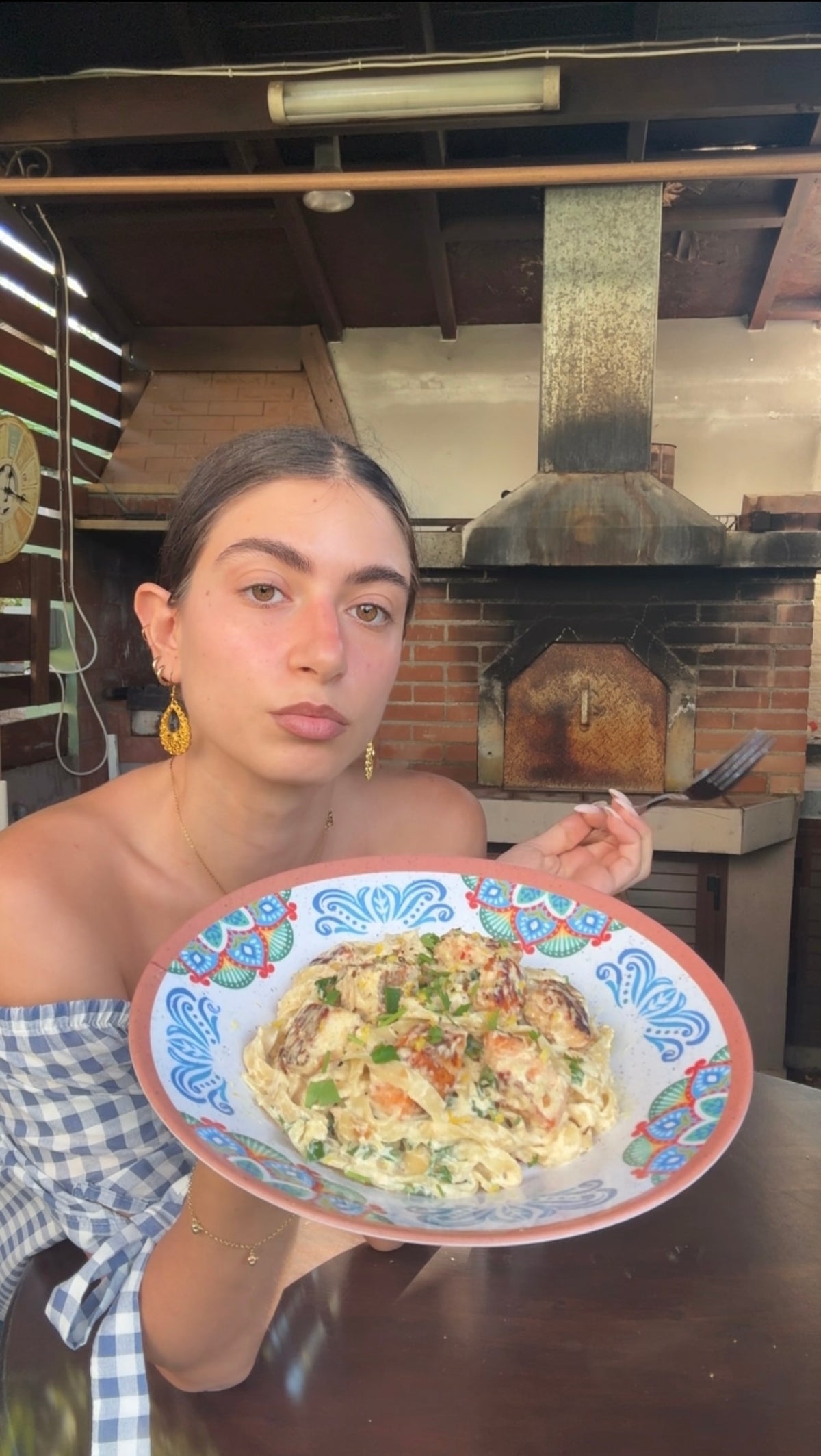 A woman in a blue-checkered dress holds a colorful plate of pasta, posing in front of a brick oven in an outdoor kitchen.