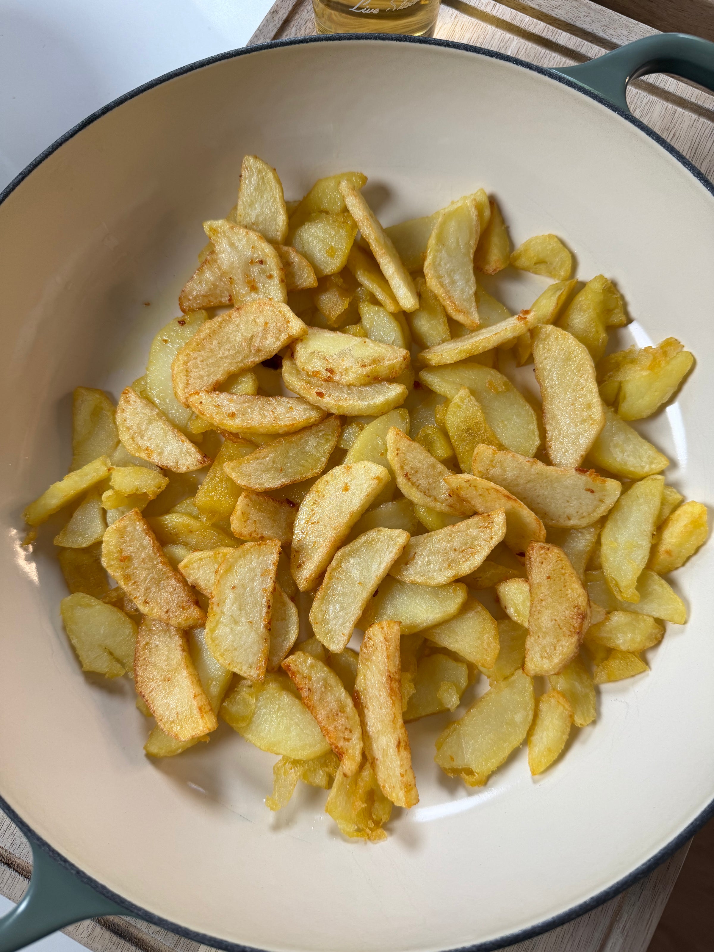 A cream-colored pan filled with golden, roasted potato wedges, some slightly crispy and seasoned, sits on a wooden surface.