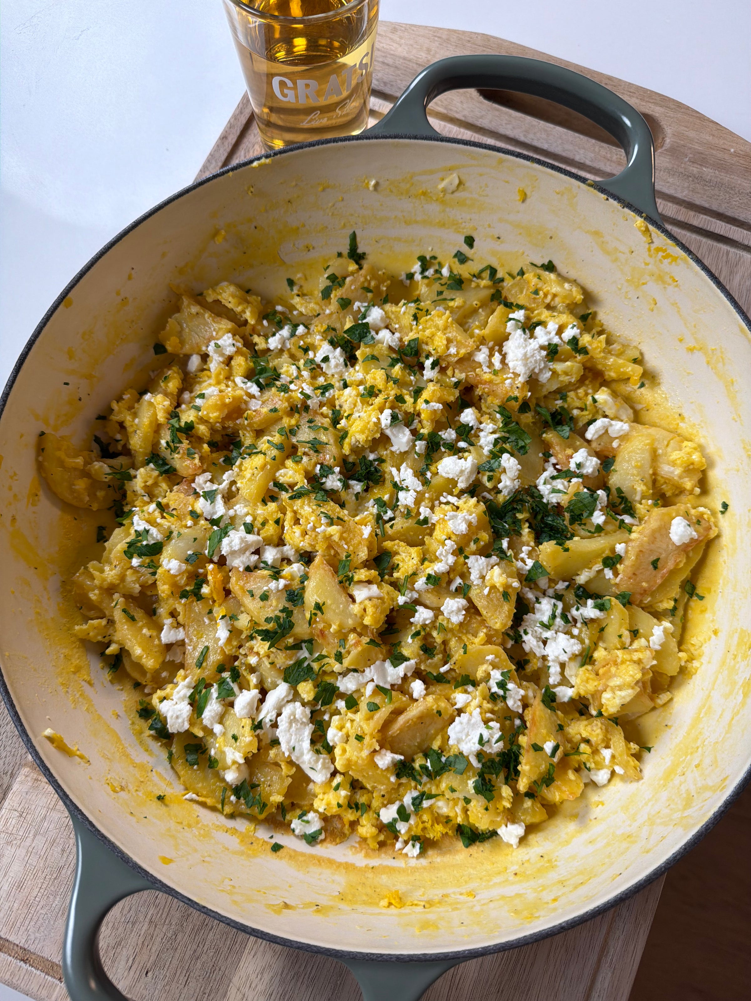A green-handled skillet filled with scrambled eggs, potato slices, chopped herbs, and crumbled white cheese sits on a wooden board. A glass of amber liquid is in the background.