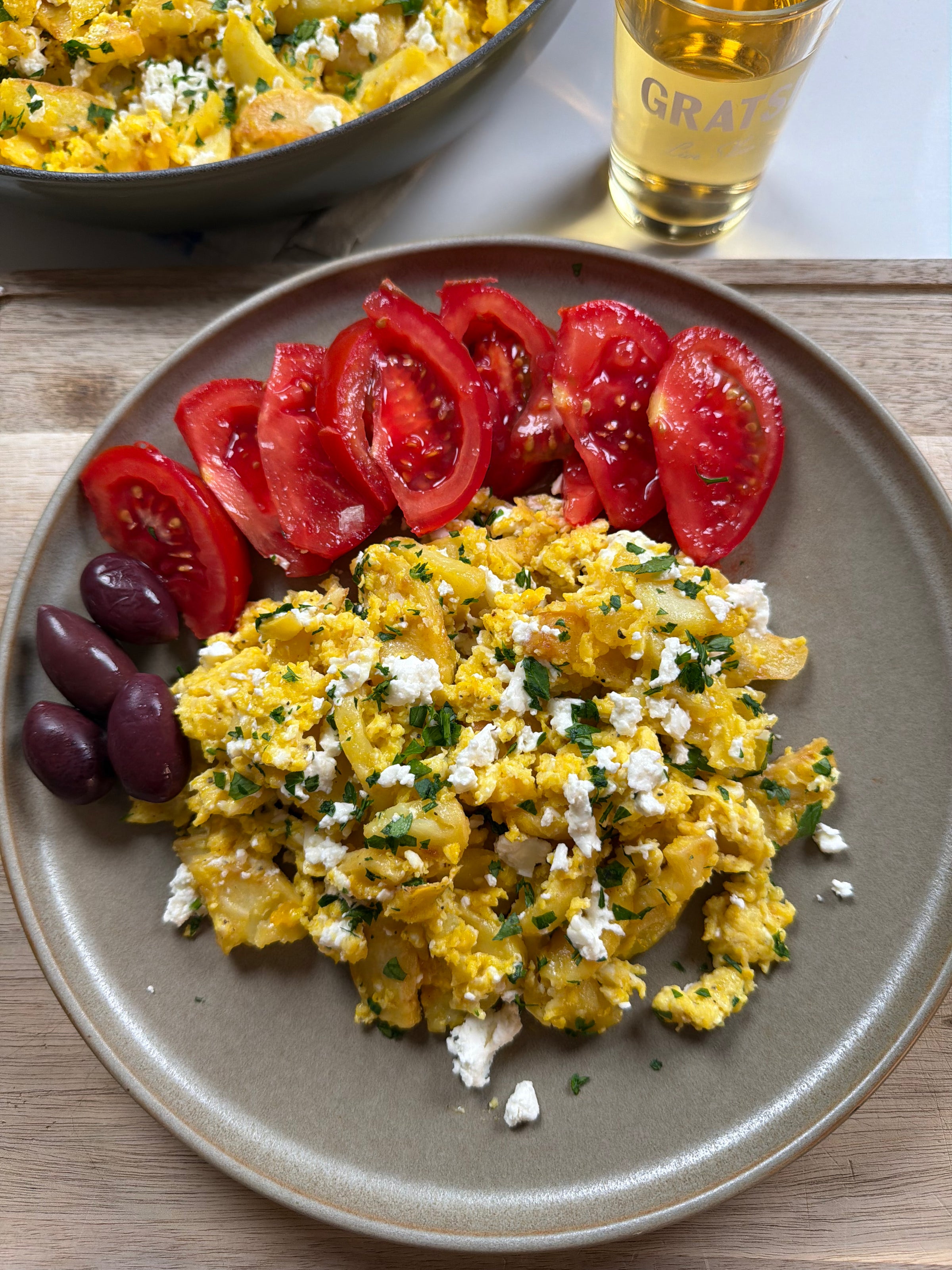 A plate with scrambled eggs topped with crumbled feta and herbs, sliced fresh tomatoes, and three Kalamata olives. A glass of light-colored beverage and a bowl of more eggs are in the background.