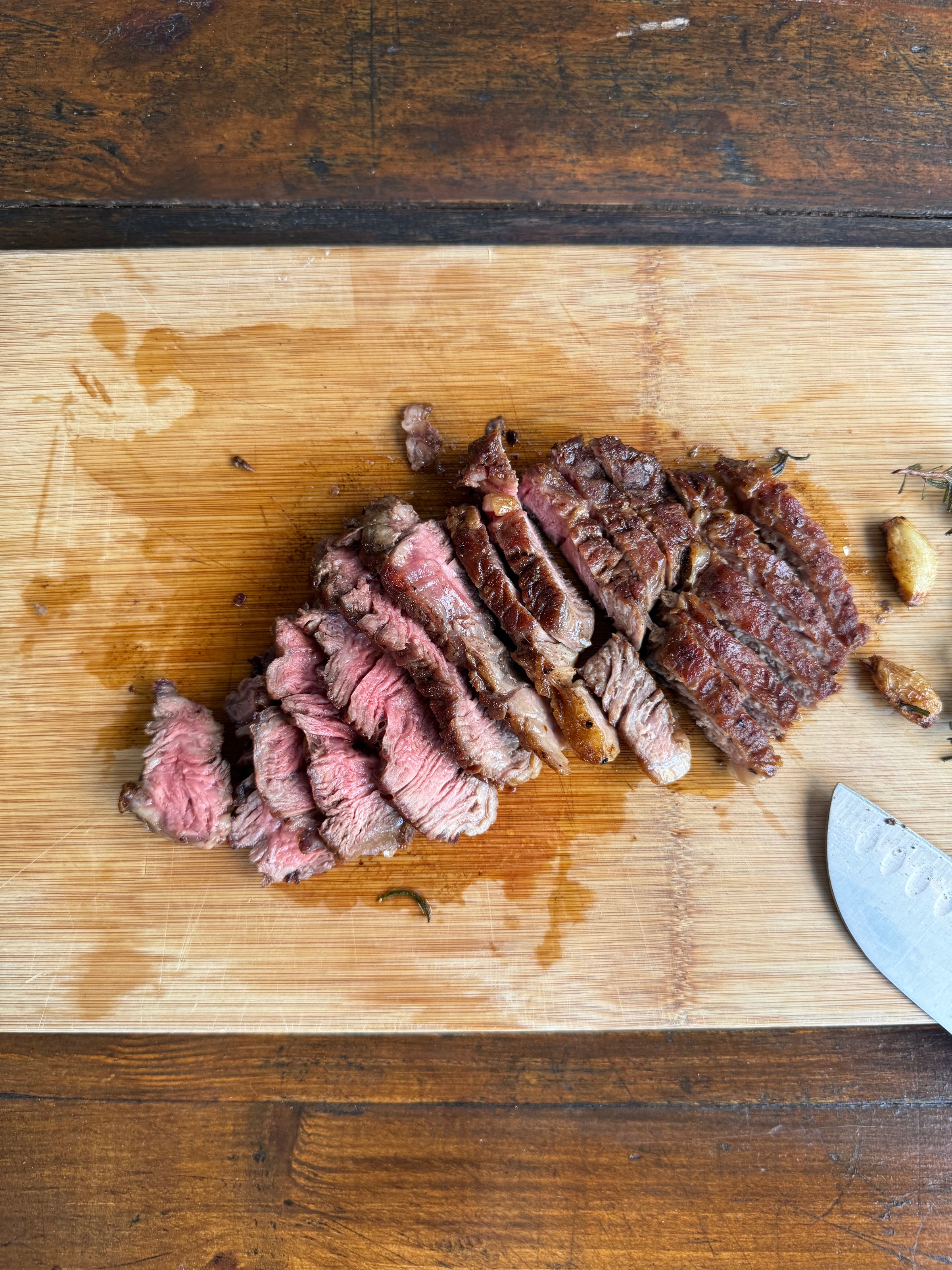 Sliced steak on a wooden cutting board with a knife and roasted garlic cloves.