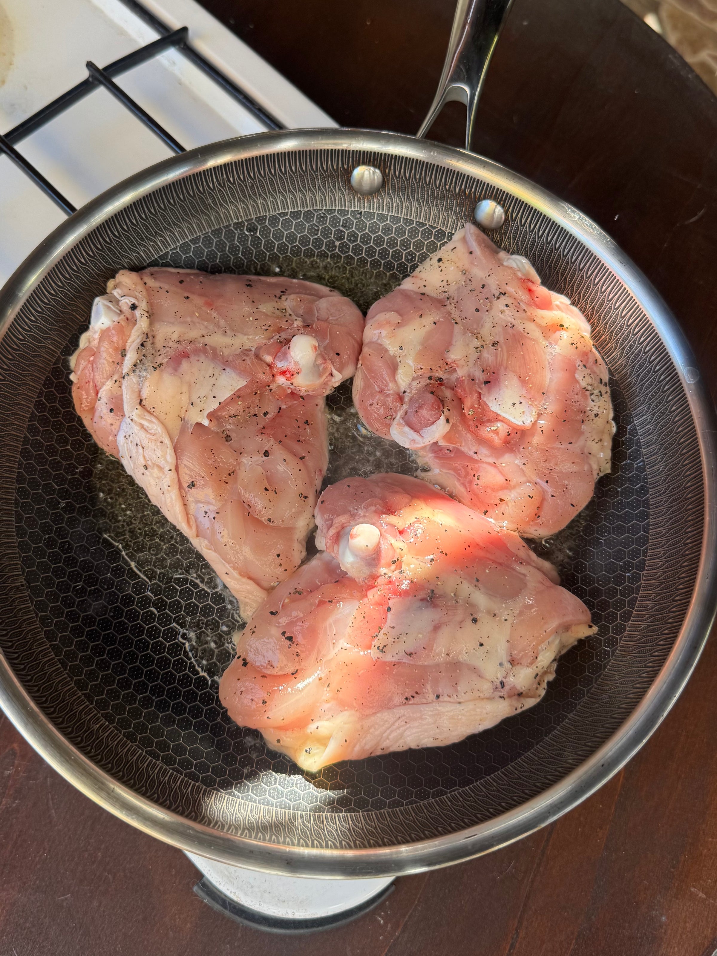 Three raw, seasoned chicken thighs are cooking in a frying pan on a stovetop, with sunlight highlighting parts of the meat.