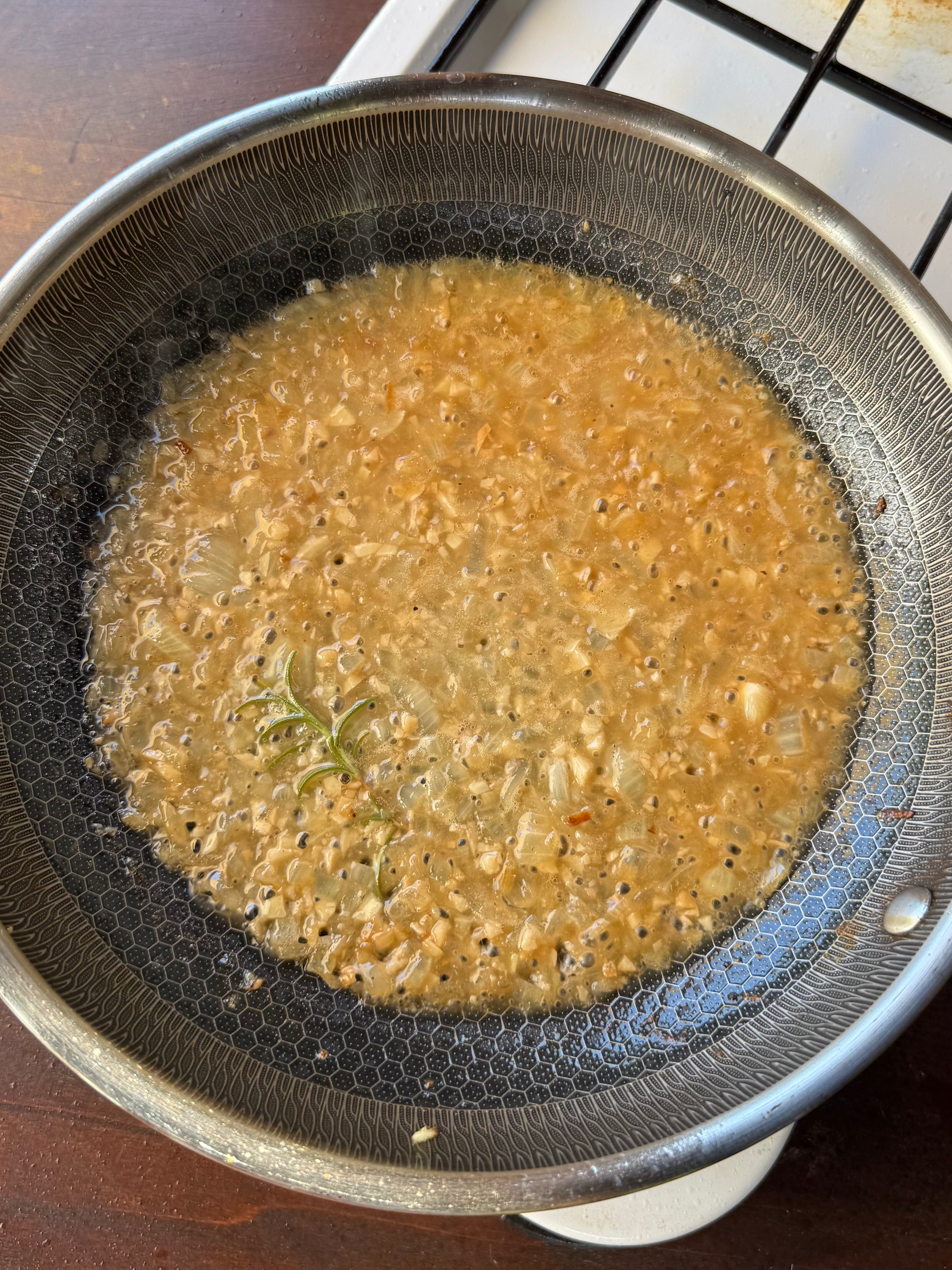 A frying pan with bubbling oil, chopped onions, mustard seeds, spices, and a sprig of fresh rosemary cooking on a stovetop.