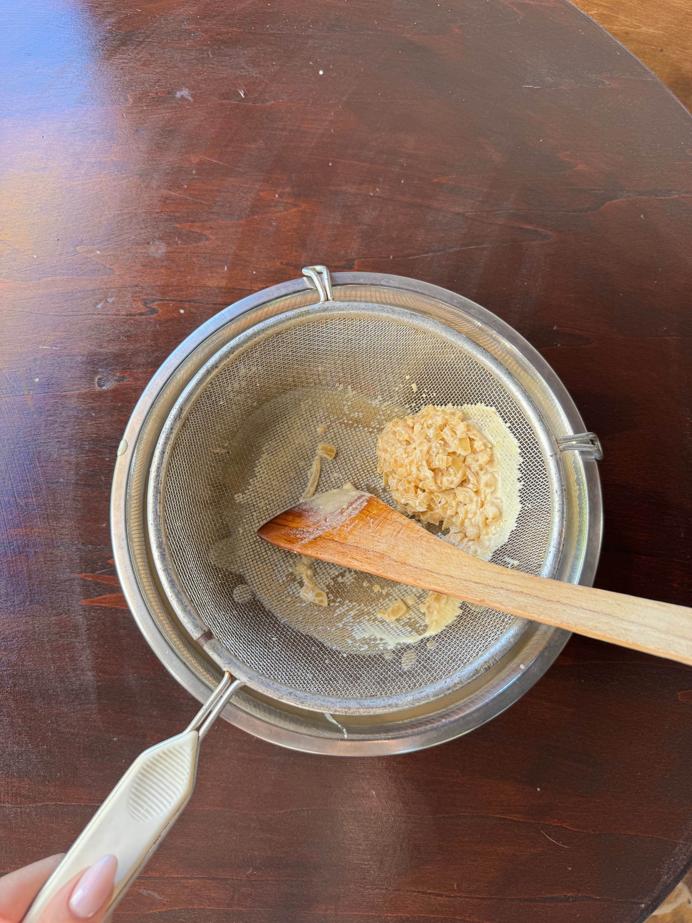 A hand holds a fine-mesh strainer over a bowl, with a wooden spoon stirring some flour and clumps left in the strainer. The surface beneath is dark wood.
