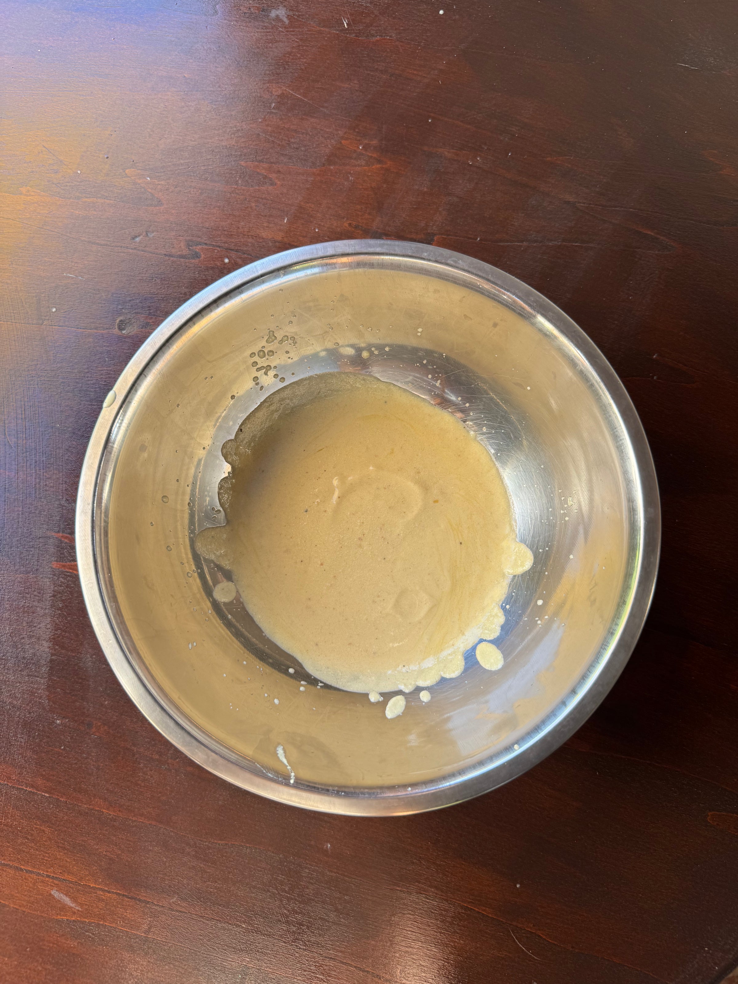 A metal bowl containing a small amount of light brown batter sits on a dark wooden surface. Some batter is splattered on the inner side of the bowl.