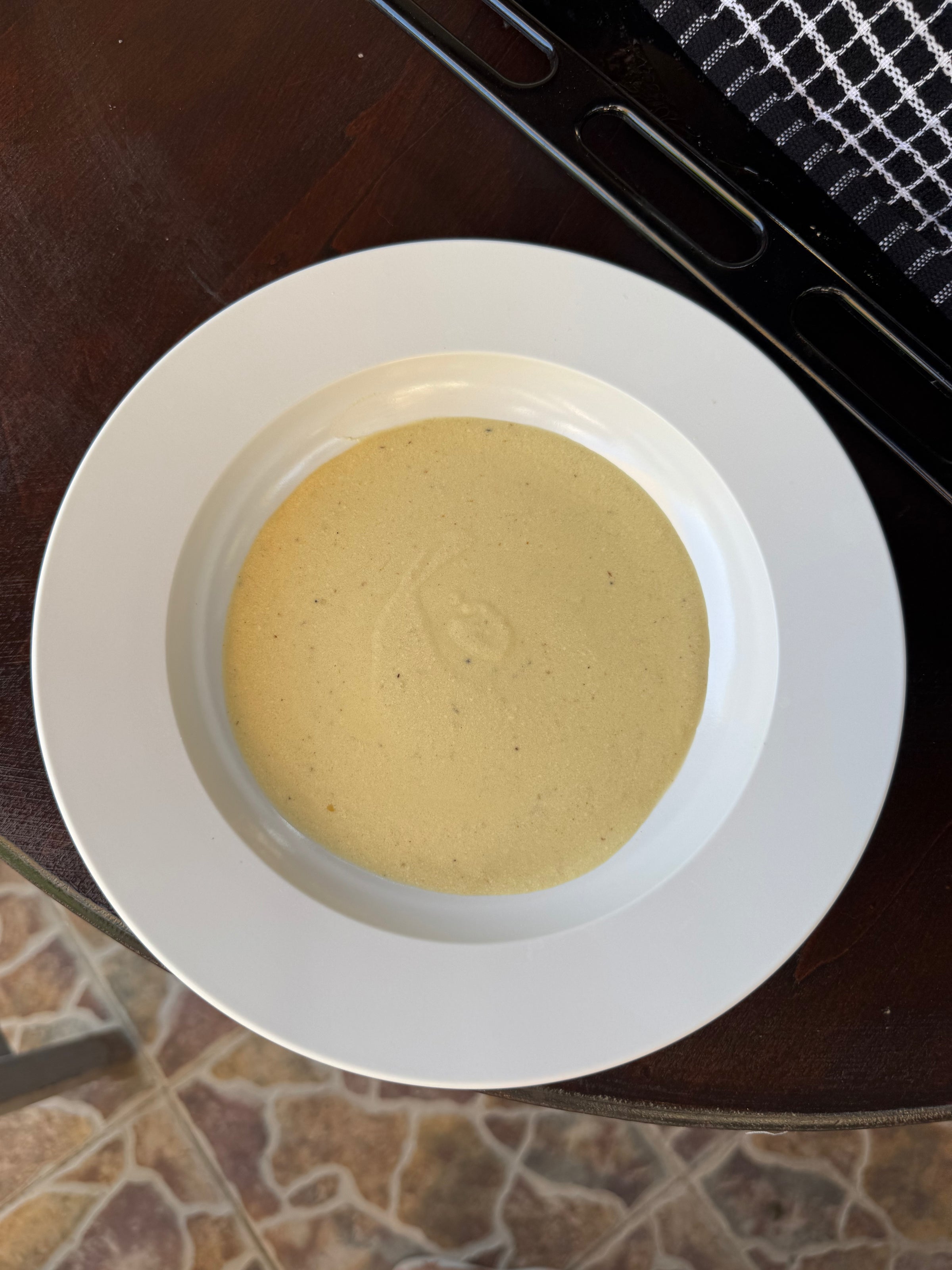 A shallow white bowl filled with a smooth, light yellow soup sits on a dark wooden table with a tiled floor visible in the background.